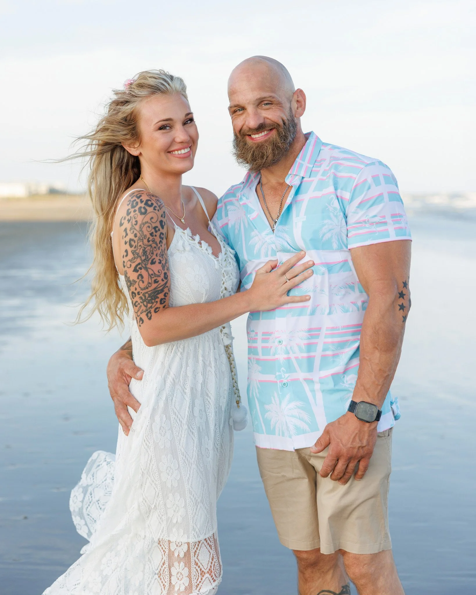 A smiling couple standing close together at the beach, with the ocean in the background. The woman has long blonde hair, a tattoo of leopard print on her arm, and is wearing a white lace dress. The man has a beard, a tattoo of stars on his arm, and i