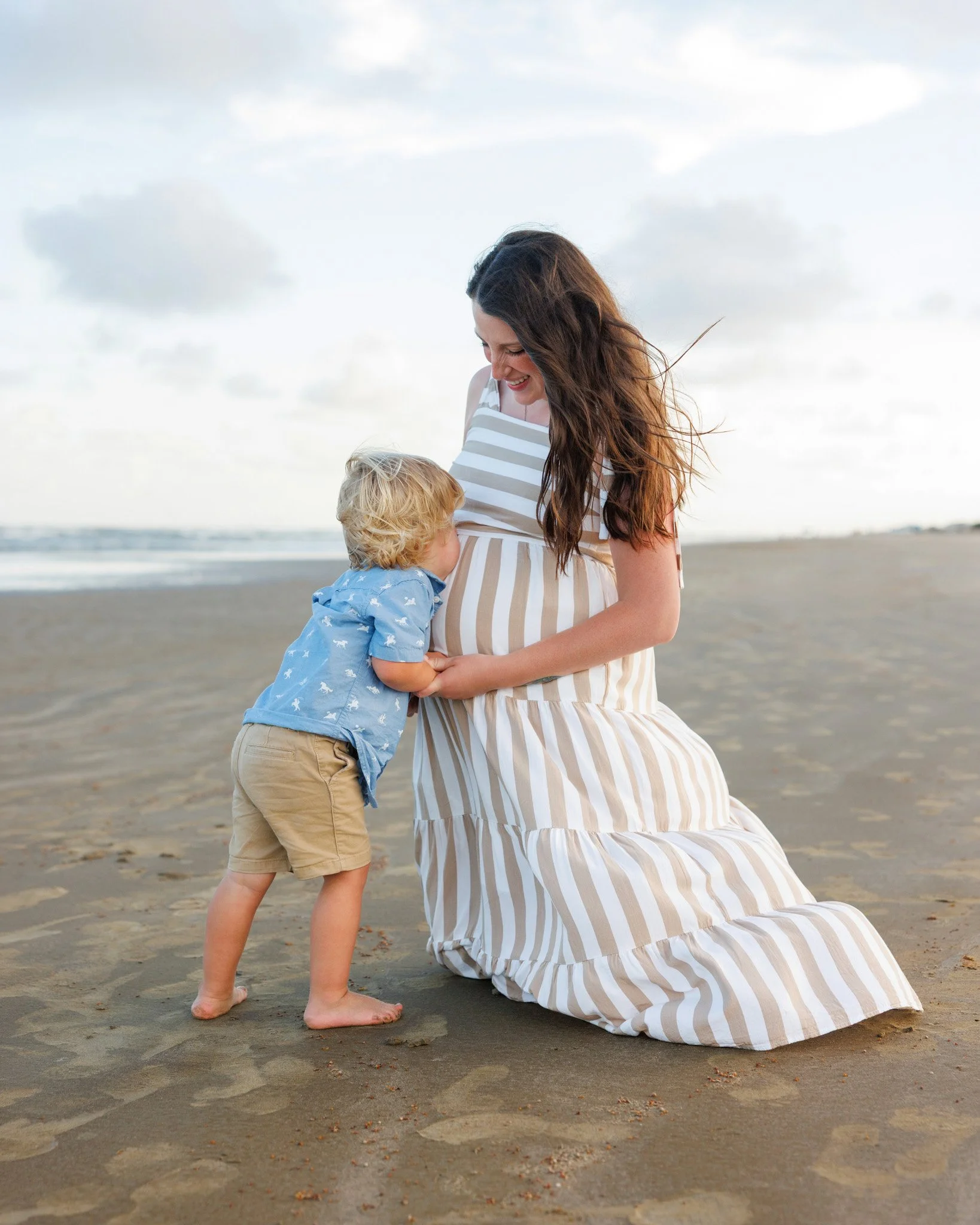 A woman and a young boy on a beach. The woman, wearing a striped dress, is kneeling and smiling at the boy. The boy, wearing a blue shirt and beige shorts, is standing barefoot on the sand, facing the woman. The sky is partly cloudy, and the ocean is