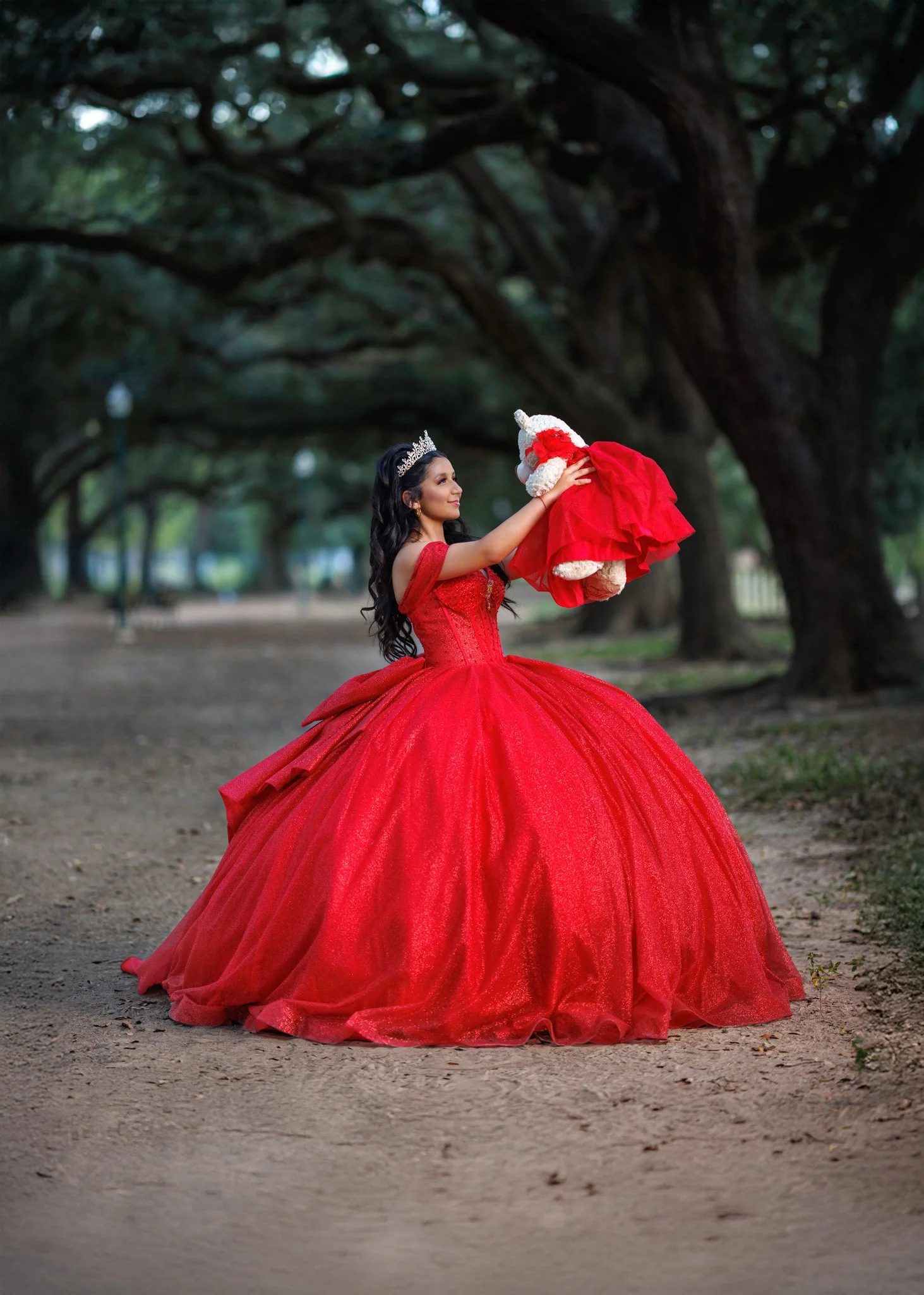 A woman in a red dress holding a teddy bear dressed in red, standing outdoors under large, twisted trees.