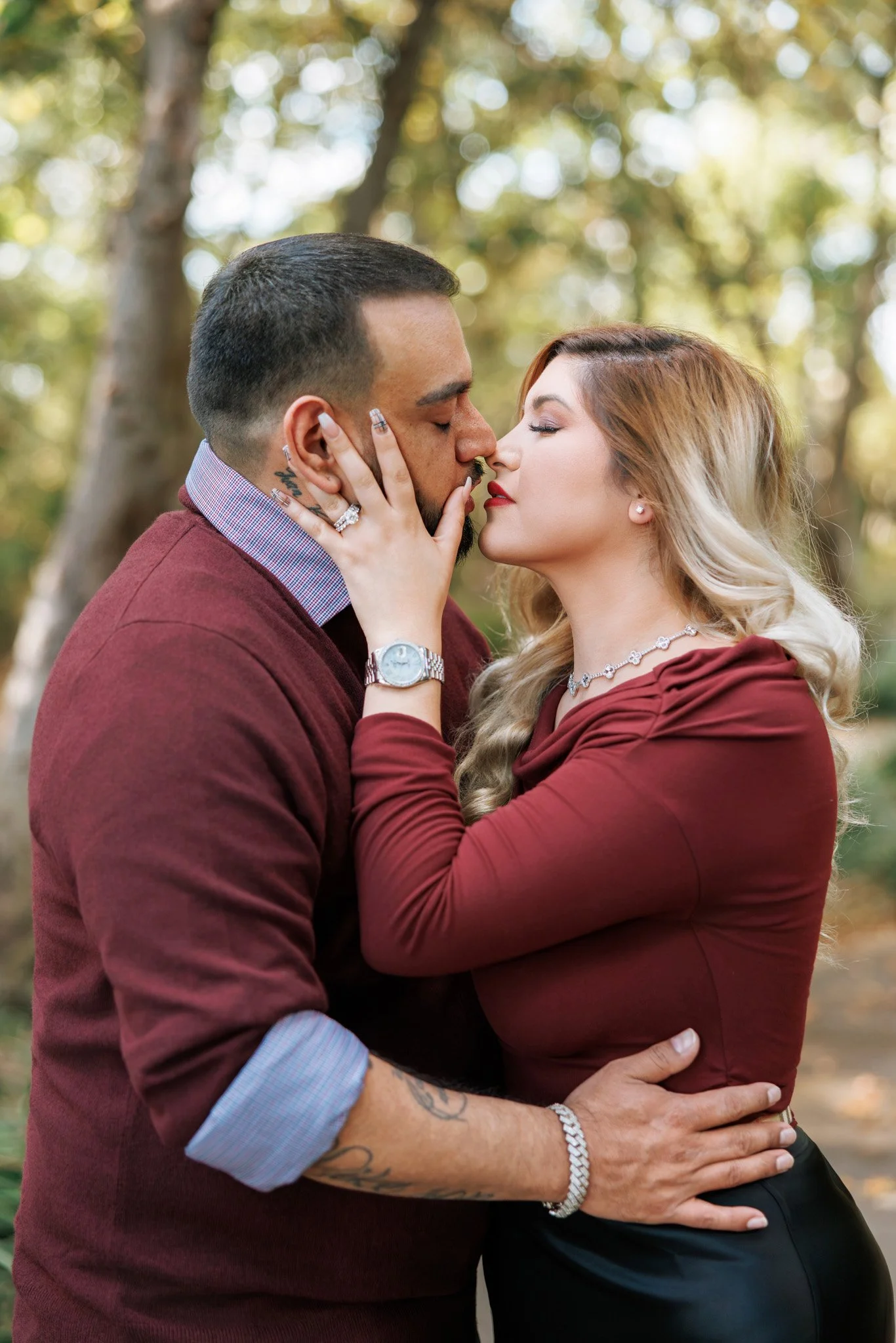 A couple passionately embraces and kisses outdoors with blurred trees in the background. The man is wearing a maroon sweater and has tattoos, while the woman has long blonde hair, red lipstick, and is dressed in a maroon top and black skirt, accessor