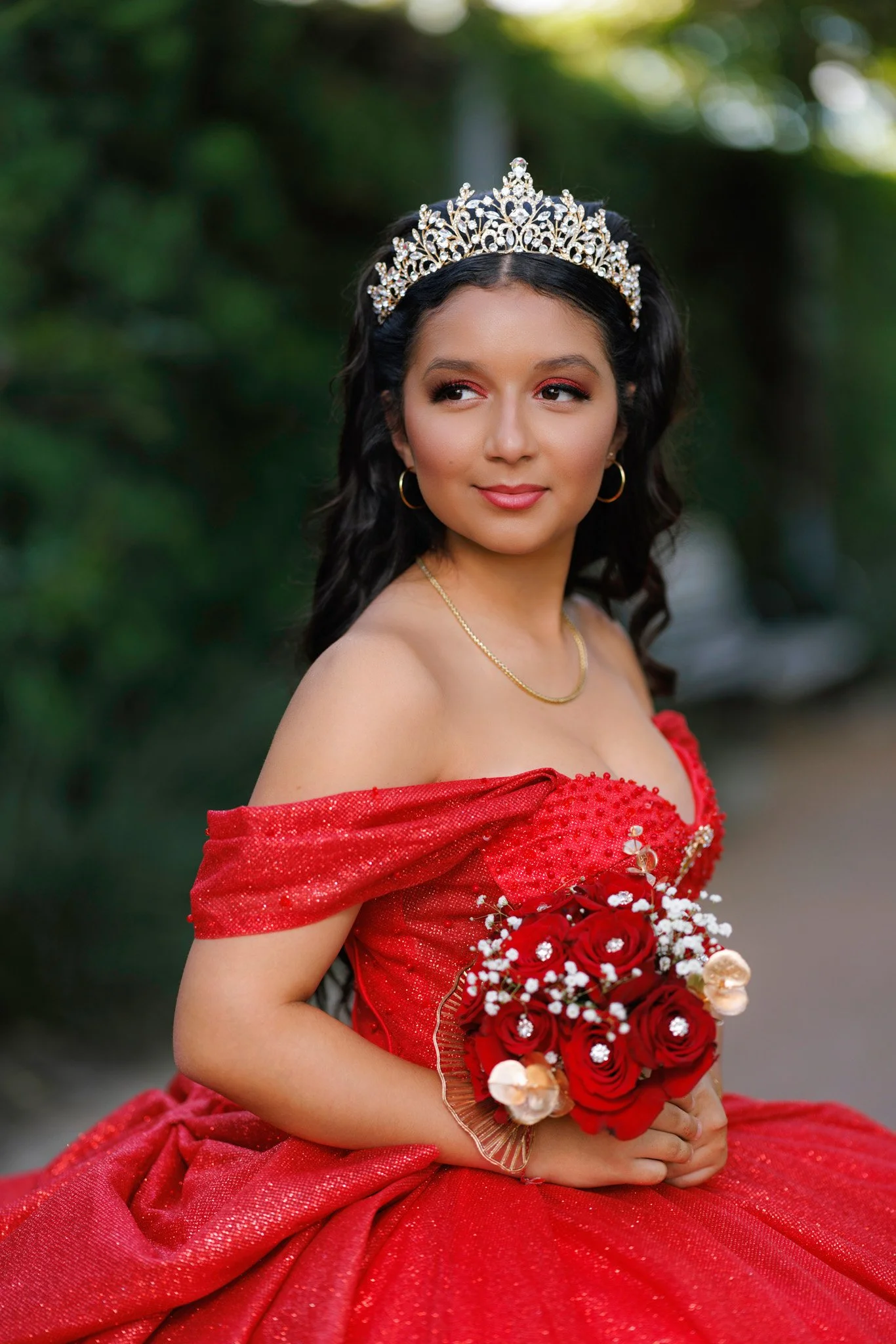 Young woman in a red gown and tiara holding a bouquet of red roses with white baby's breath, outdoors with blurred green foliage background.