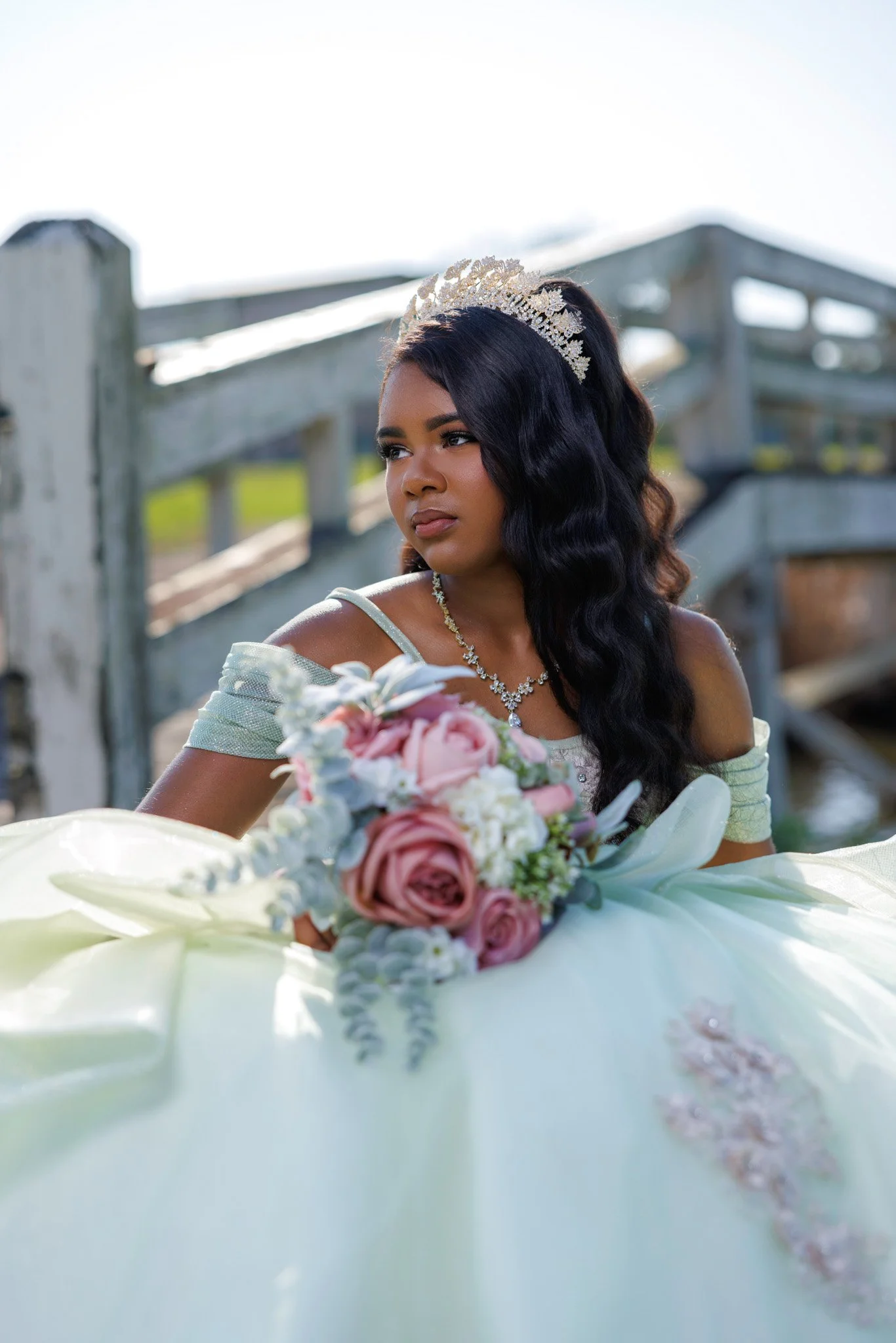 A young woman with long, dark, wavy hair wearing a tiara and jewelry, holding a bouquet of pink and white flowers outdoors near a wooden fence, during daytime.