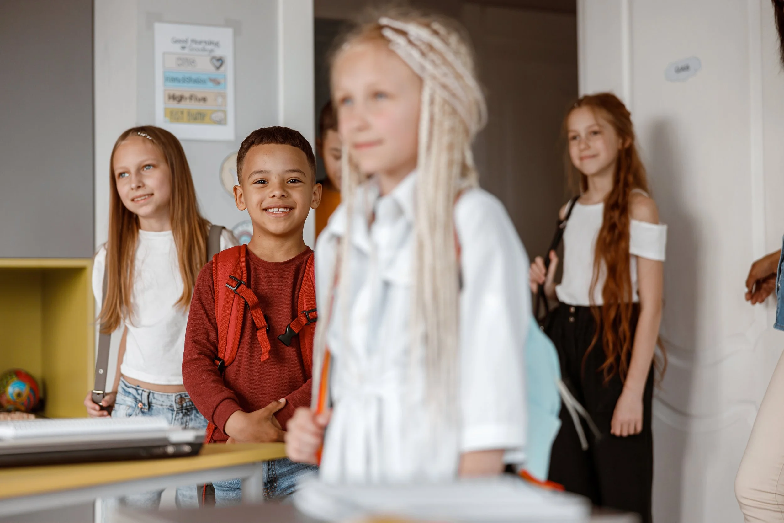 Group of children with backpacks standing in a classroom, smiling and looking towards the camera.
