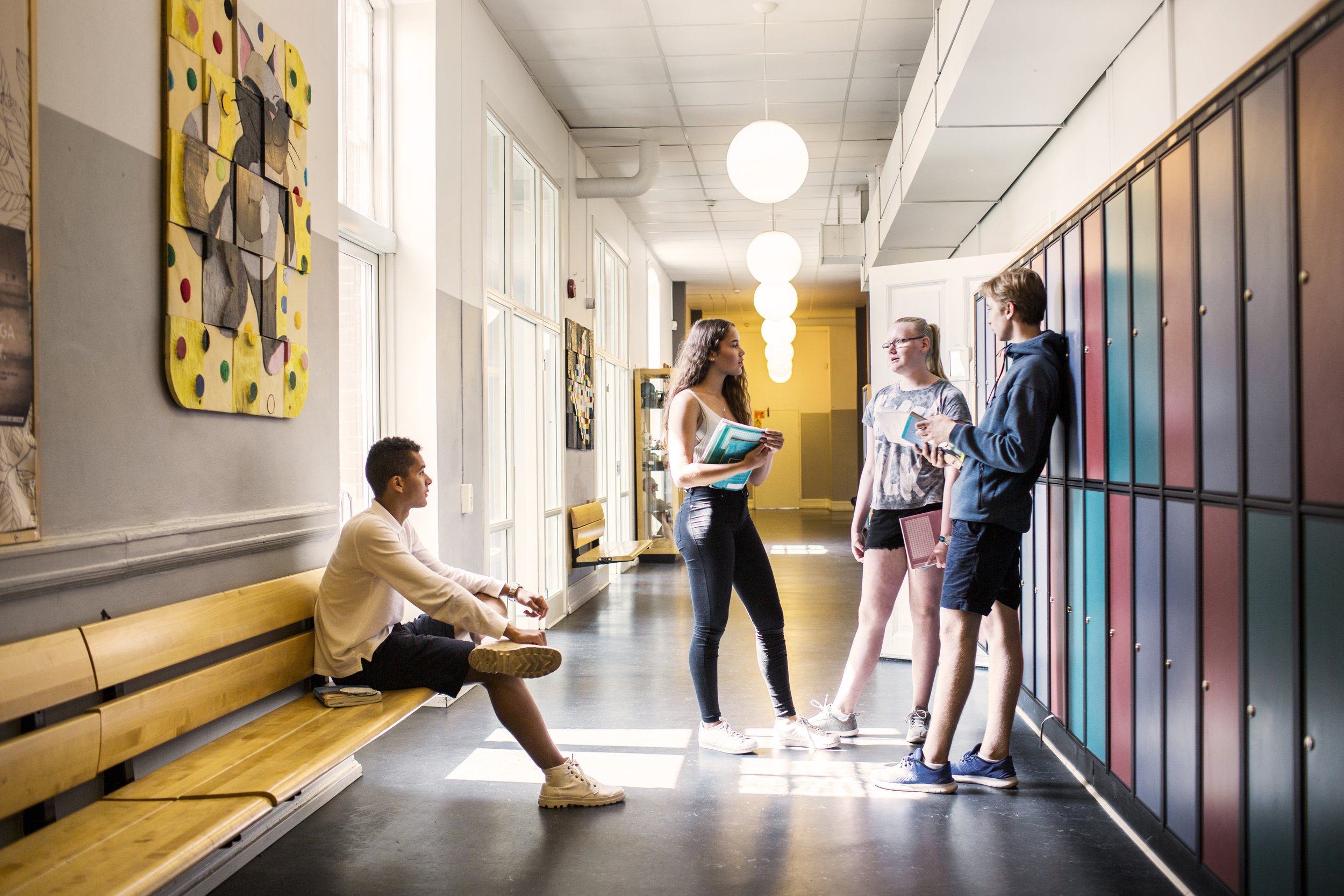 Four students in a school hallway, three standing by lockers and one sitting on a bench.