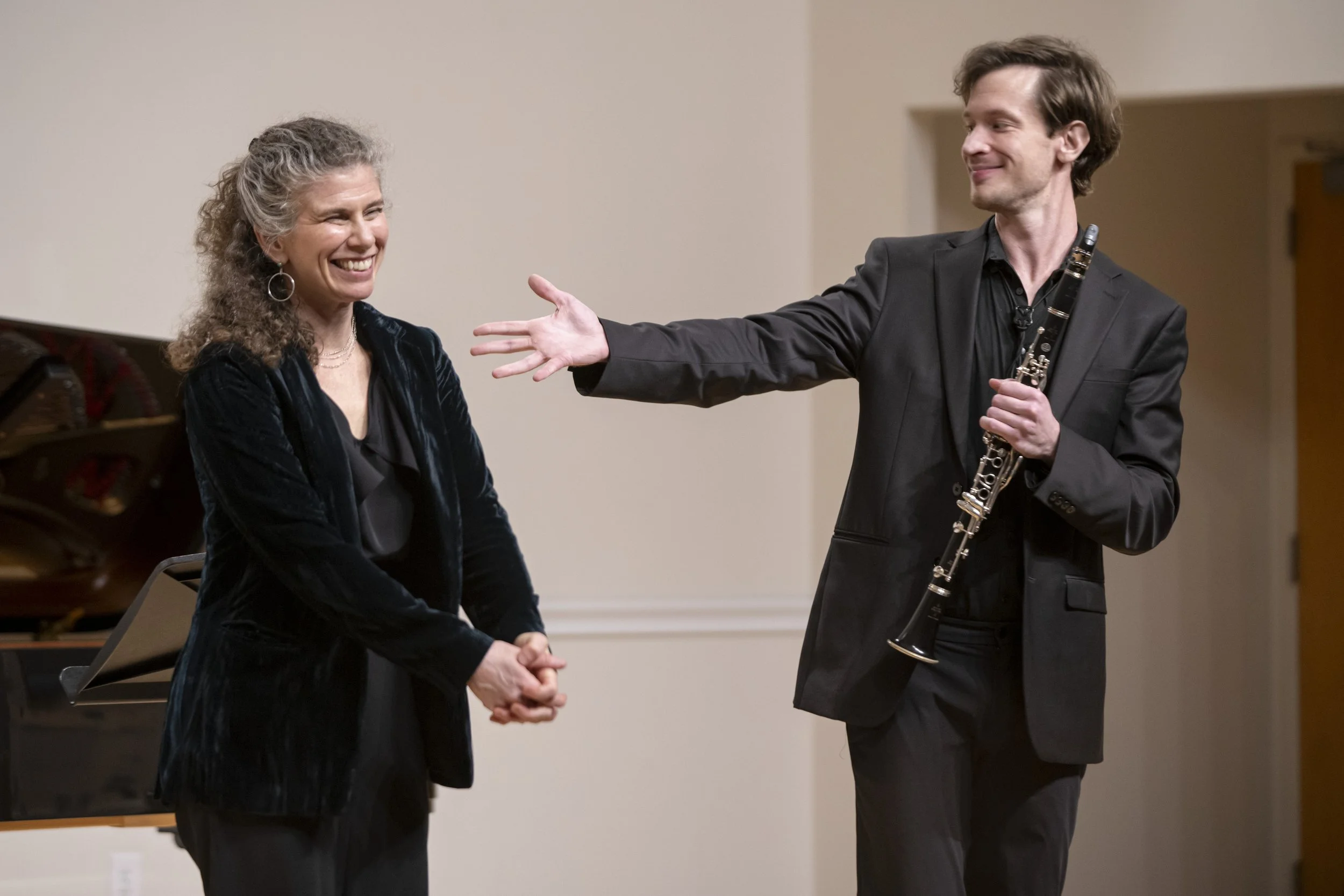 A man holding a clarinet and extending his hand towards a smiling woman in a black velvet blazer, in a music performance setting.
