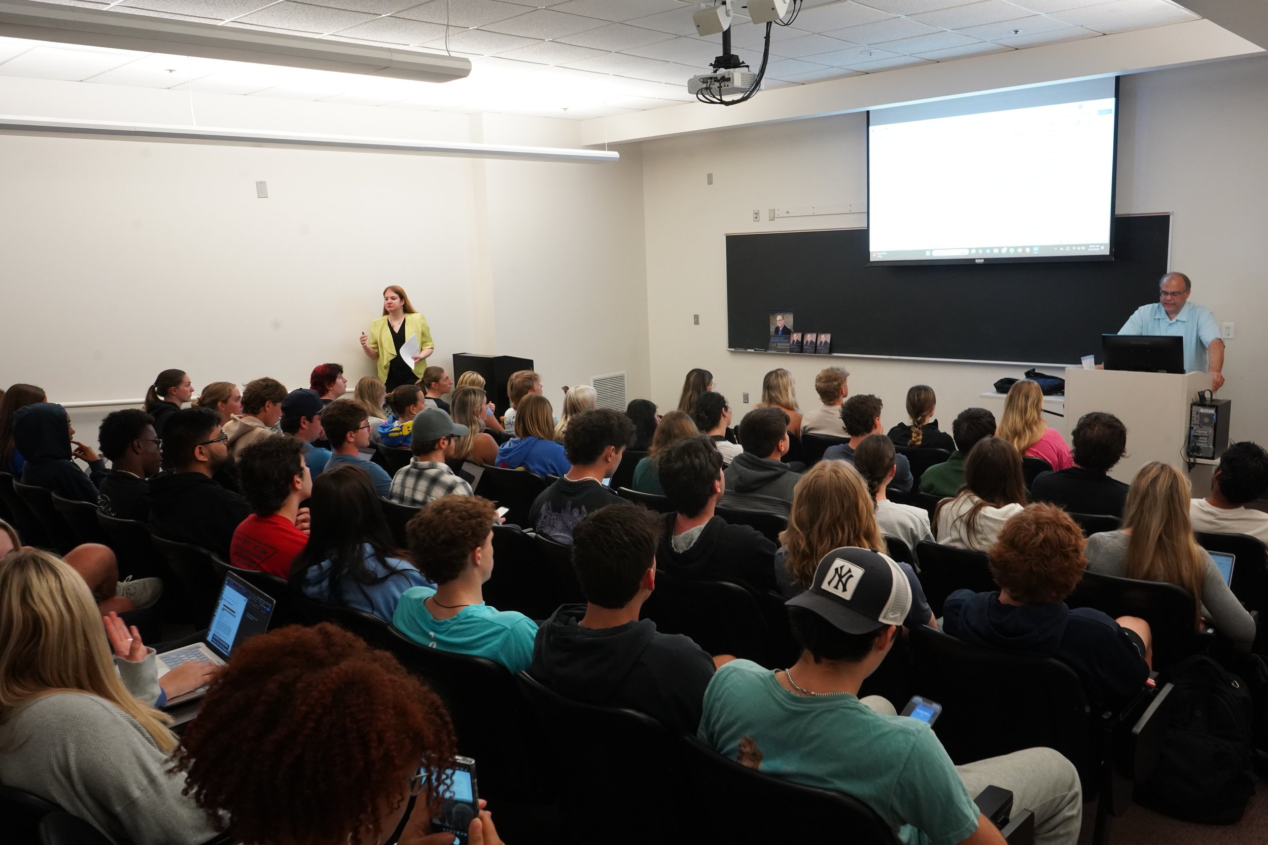 A large classroom filled with students seated and listening to a lecture, with a lecturer at a podium and a woman standing at the front.