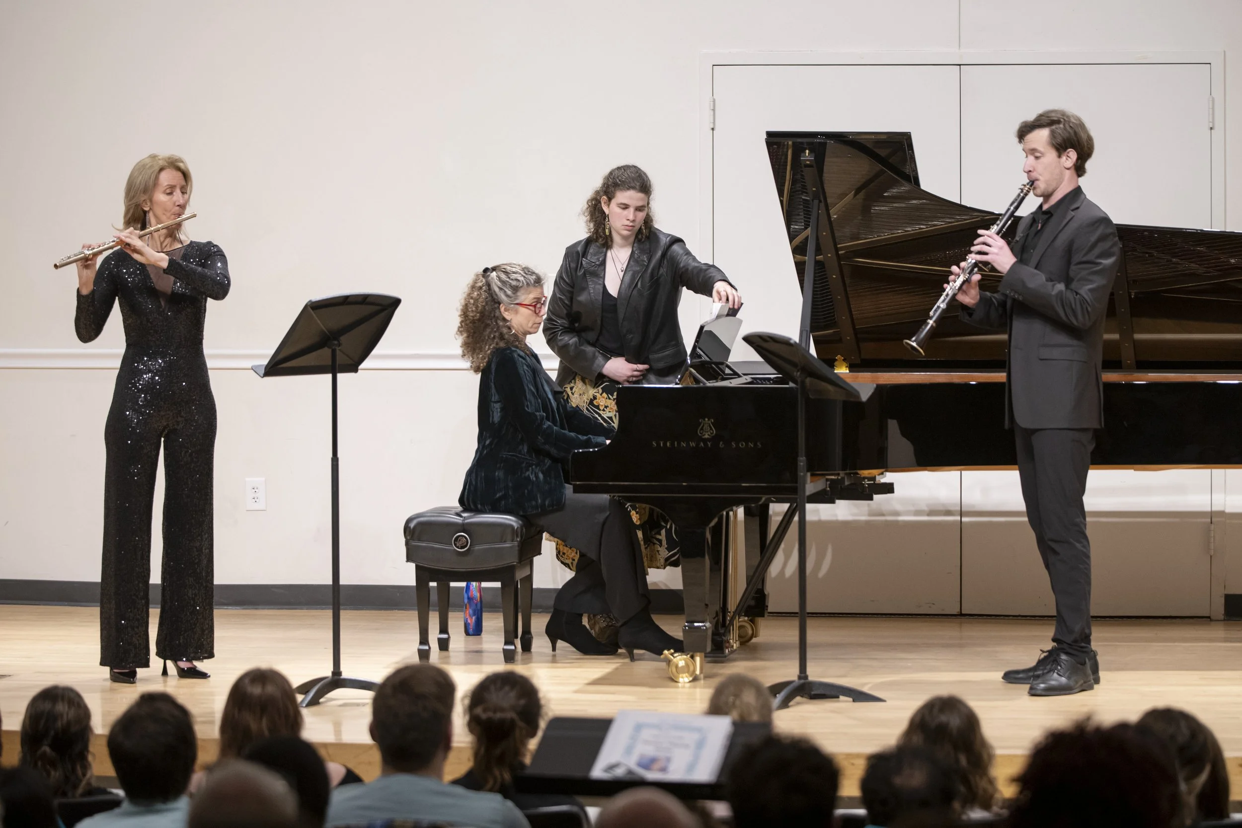 Musicians performing on stage includes a woman playing flute, a woman at piano, a woman adjusting sheet music, and a man playing clarinet. Audience seated in front.