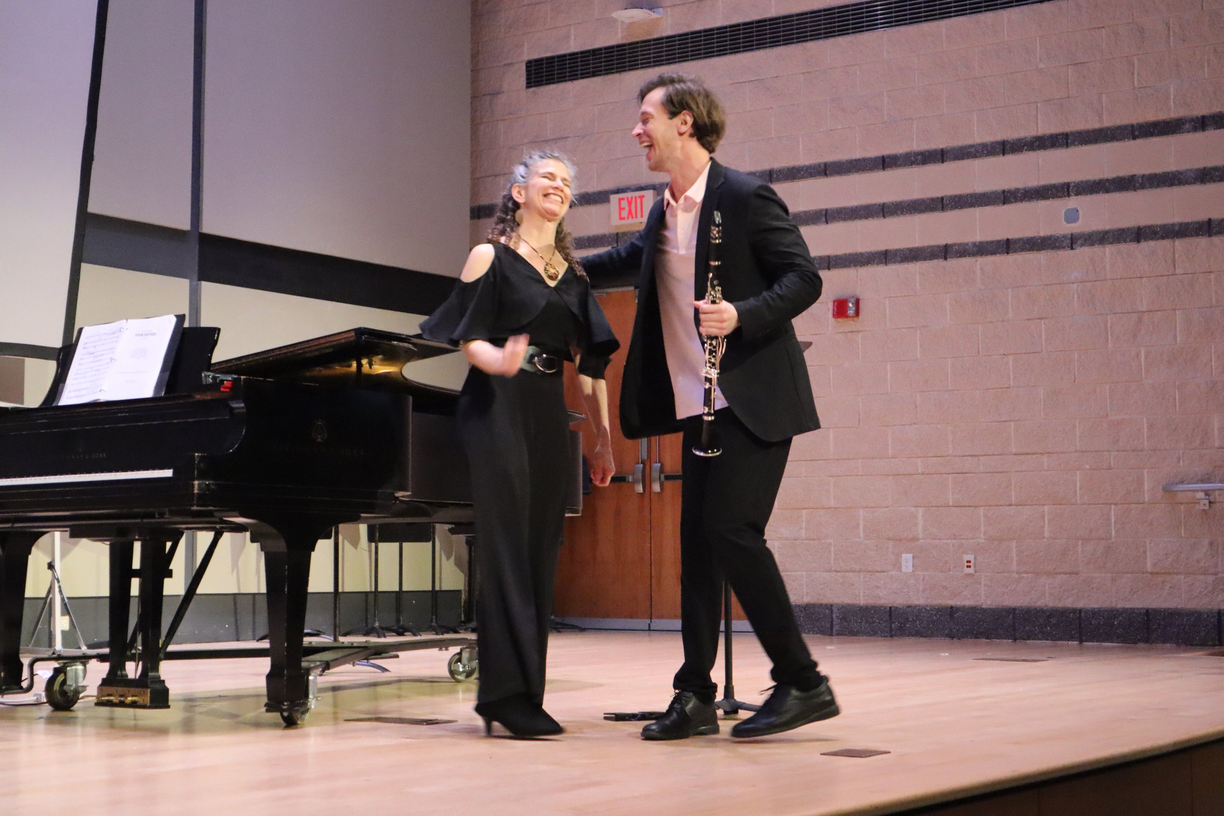 Two musicians, a woman and a man, are on a stage, smiling and looking at each other. The woman stands near a black grand piano, and the man holds a clarinet. The background is a brick wall with an exit sign.