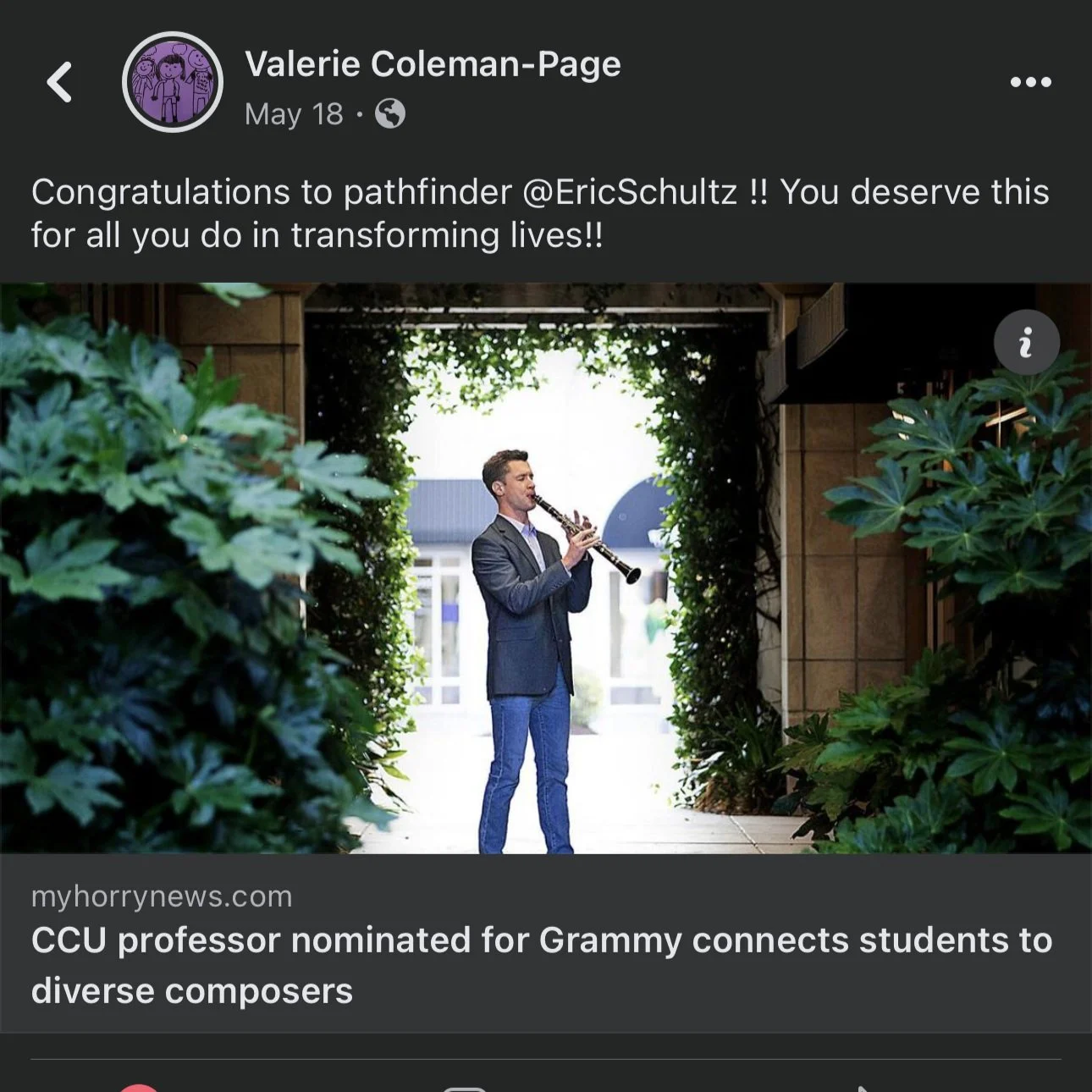 Man playing a clarinet in an outdoor setting, framed by greenery and trees, with a building in the background.