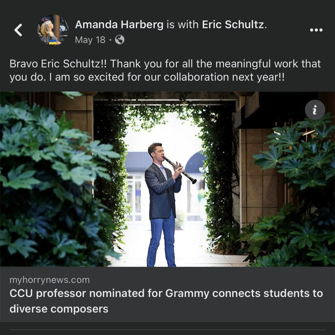 A man in a suit playing the clarinet outdoors in an archway surrounded by greenery, with a house and trees in the background.