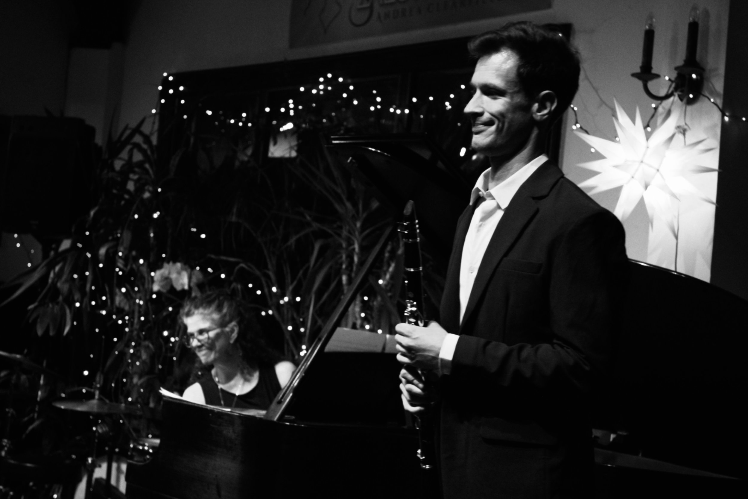 A man in a suit holding a clarinet, smiling with a woman playing the piano in the background. The scene is decorated with string lights and a large star-shaped ornament on the wall.