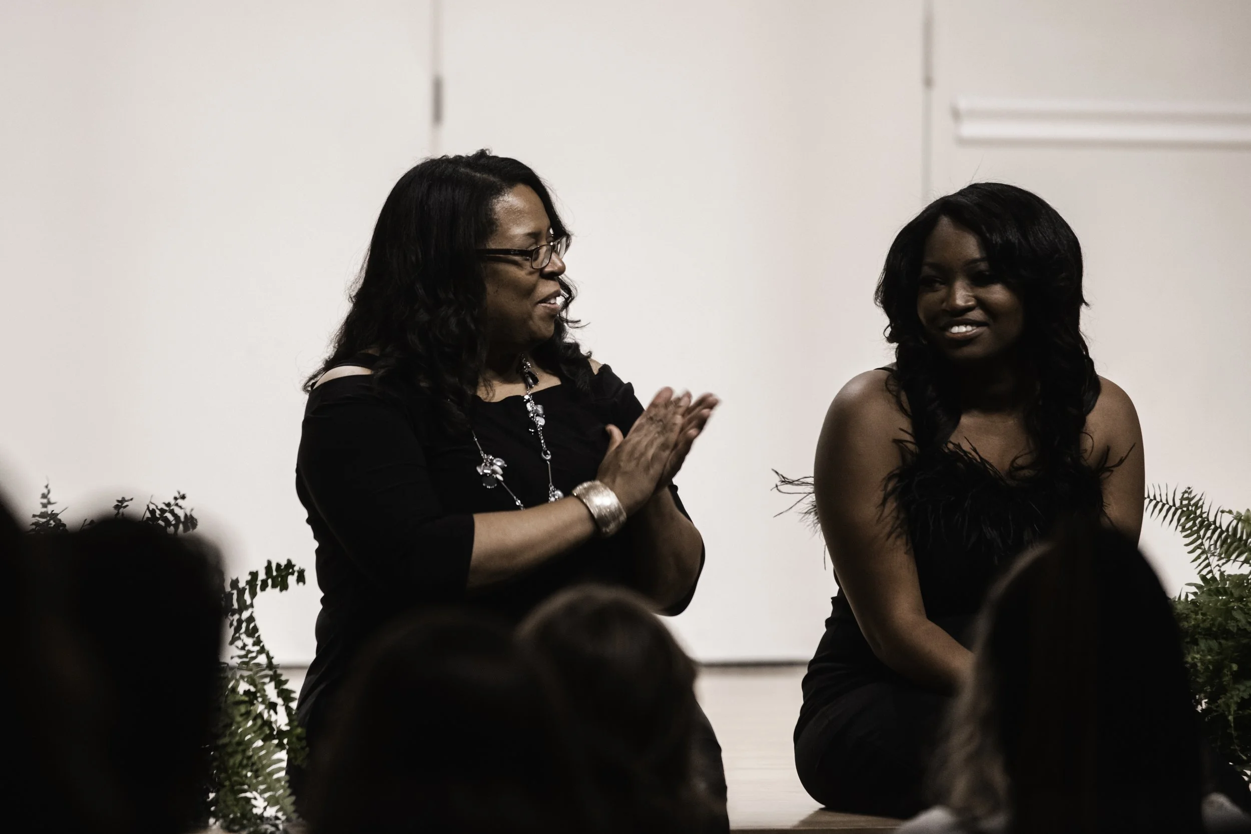 Two women are sitting on a stage, facing each other, engaged in conversation. One woman is wearing glasses and a black dress with jewelry, while the other woman has long black hair and is wearing a black dress with feather details on the top.
