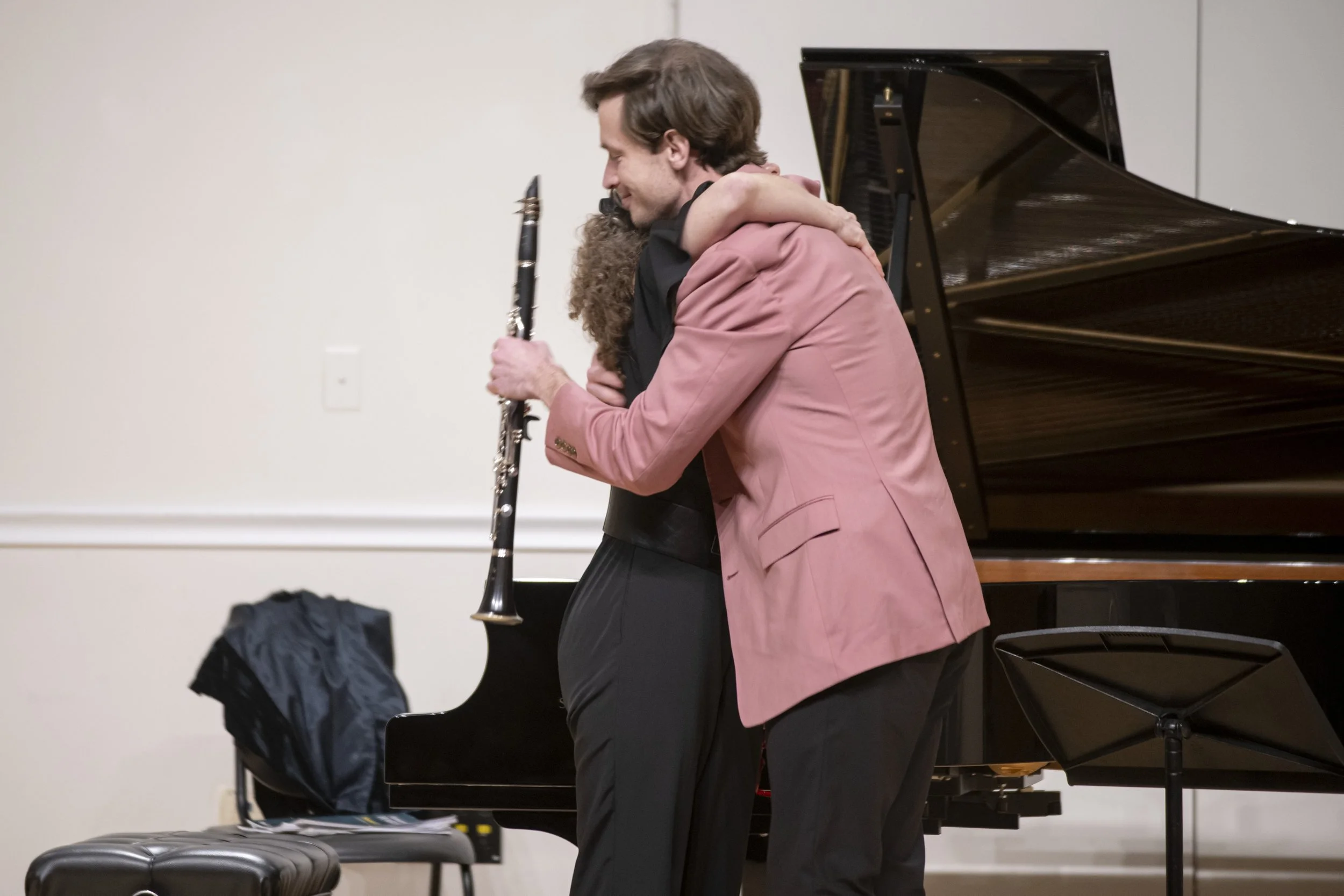 Two men embrace each other on stage, one holding a clarinet, with a grand piano in the background.