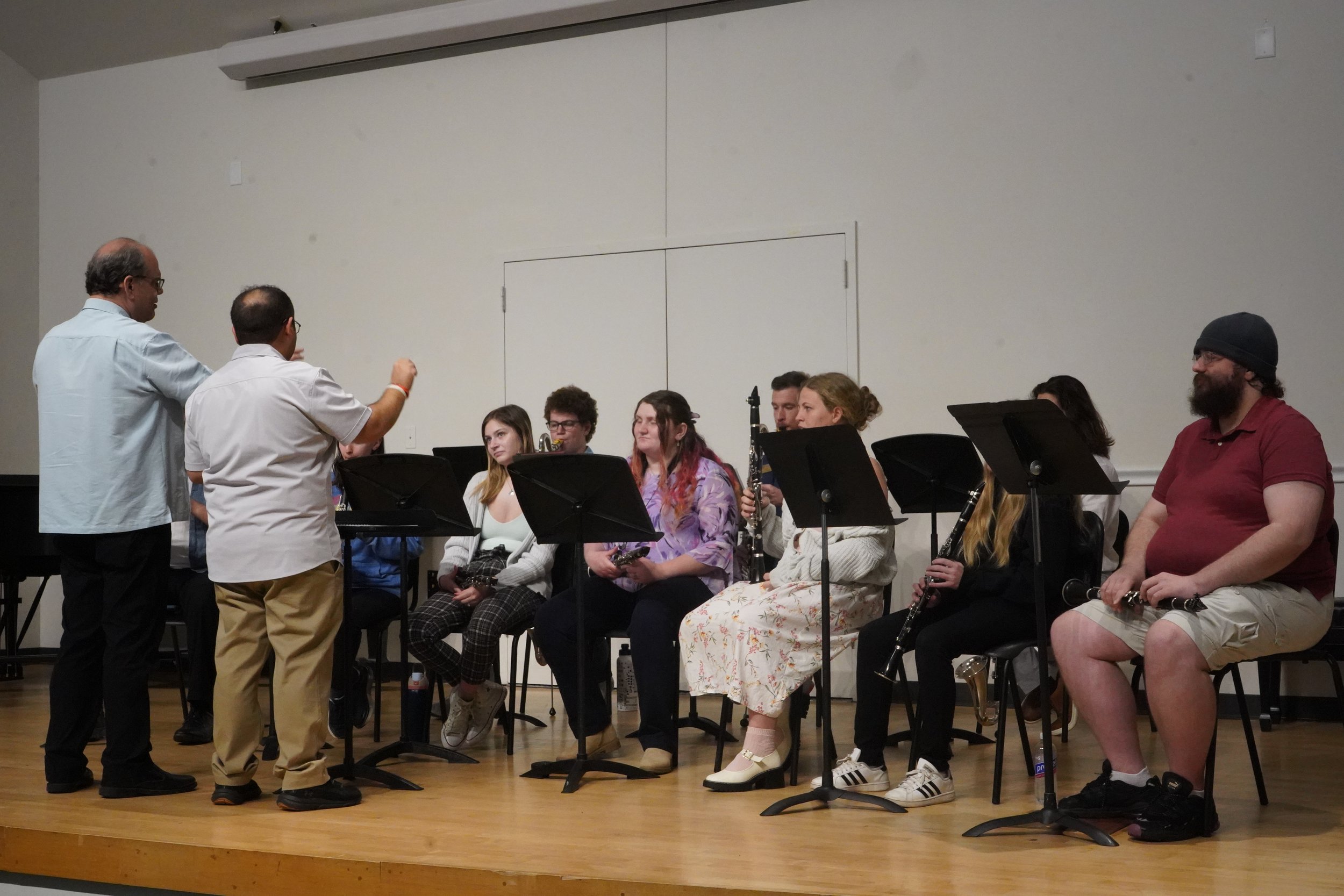 A group of people sitting on a stage, holding musical instruments, being directed by two men standing in front of them, in a room with a white wall.