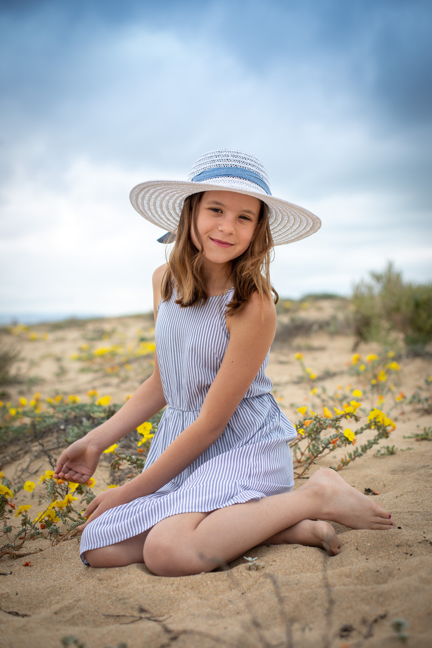 Girl-sitting-sand-yellow-flowers-white-hat-blue-ribbon.jpg