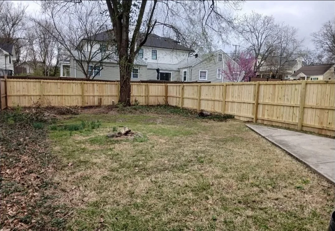 A backyard with a wooden fence, a grassy lawn, a tree, and neighboring houses in the background.