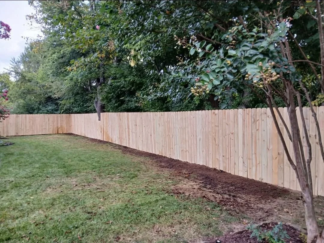 A backyard with a new wooden fence and green grass, trees, and bushes in the background.