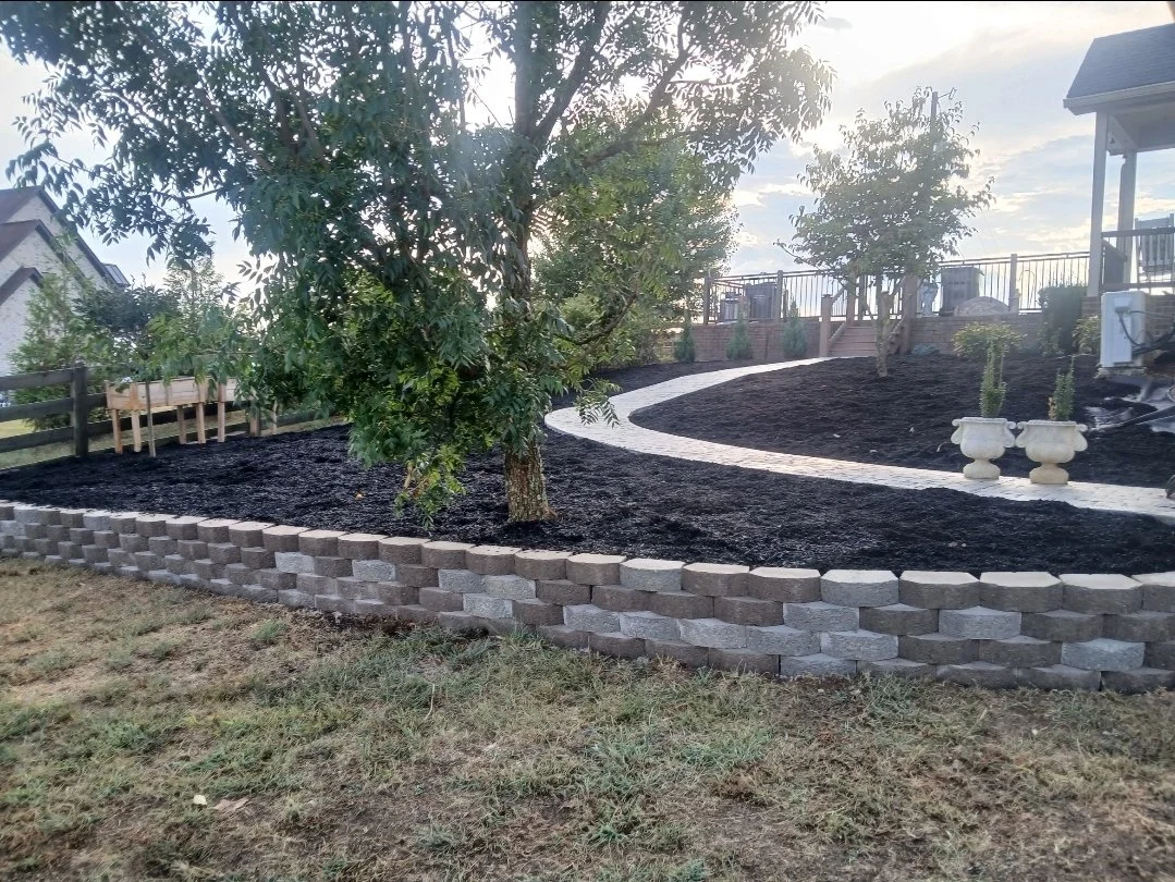 A landscaped backyard featuring a winding stone pathway, trees, potted plants, a fenced patio, and a retaining wall made of interlocking concrete blocks.