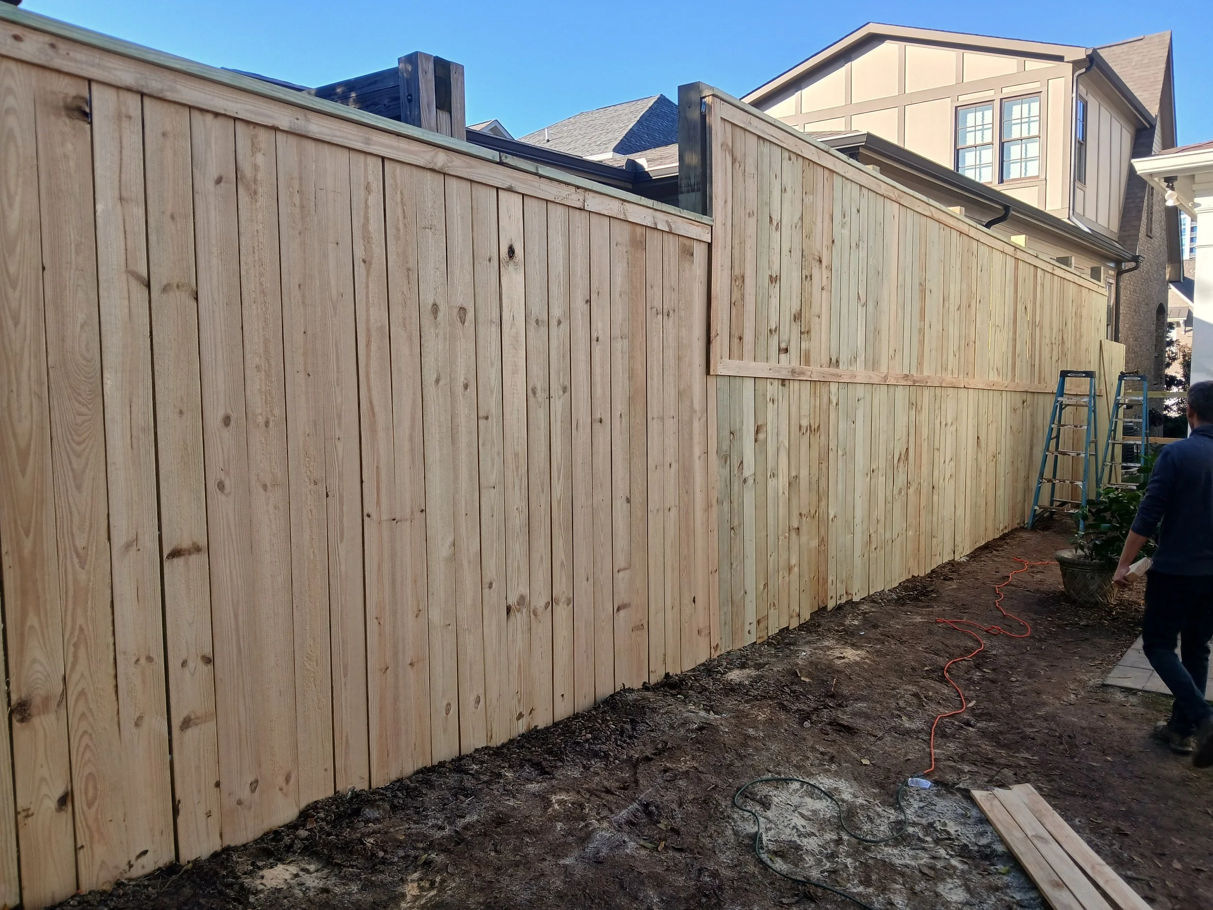 Newly built wooden privacy fence in a backyard with ladders, a person, and construction tools visible.