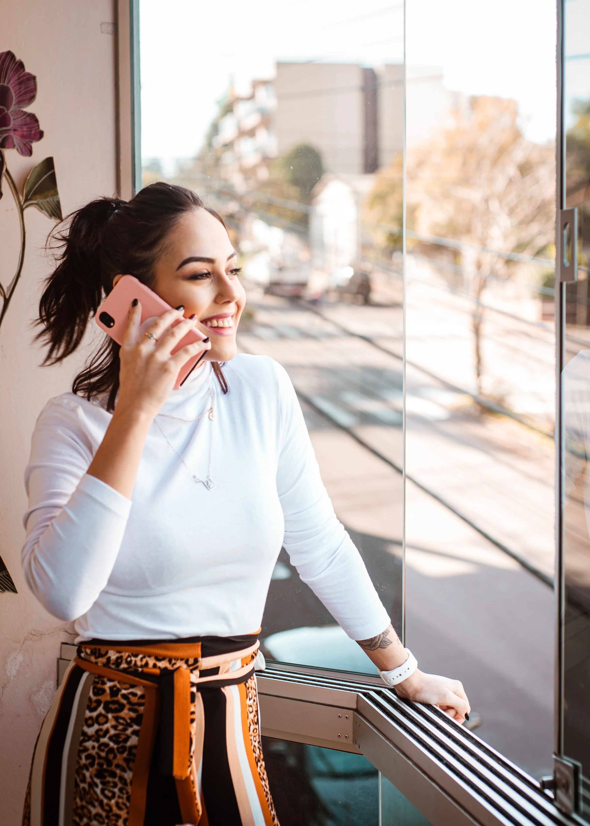 A young woman with dark hair in a ponytail, smiling and talking on a pink phone. She is wearing a white long-sleeve top, leopard print pants with a belt, and standing near a glass window with an outdoor view.