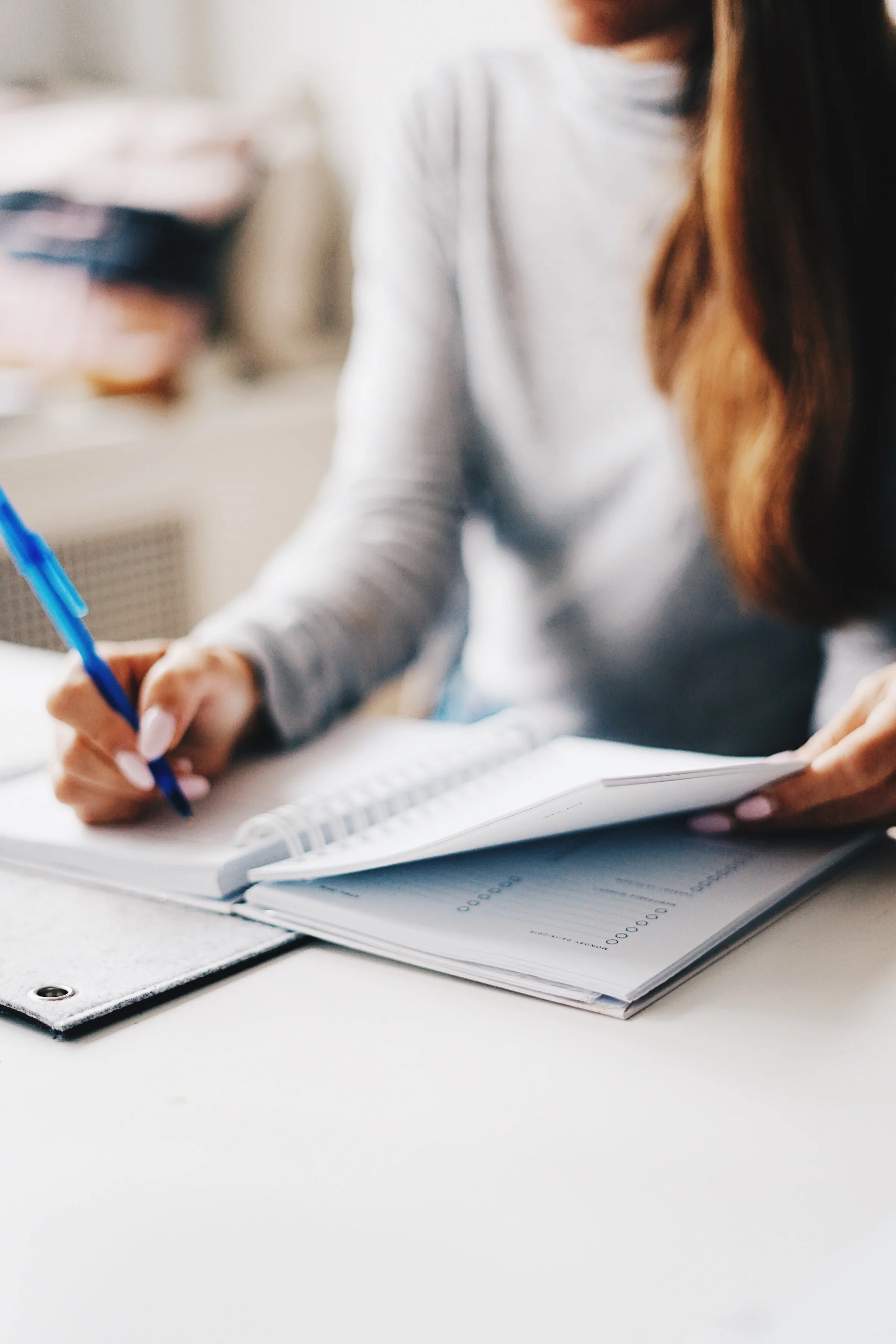 Person writing in a notebook with a blue pen, sitting at a white desk.