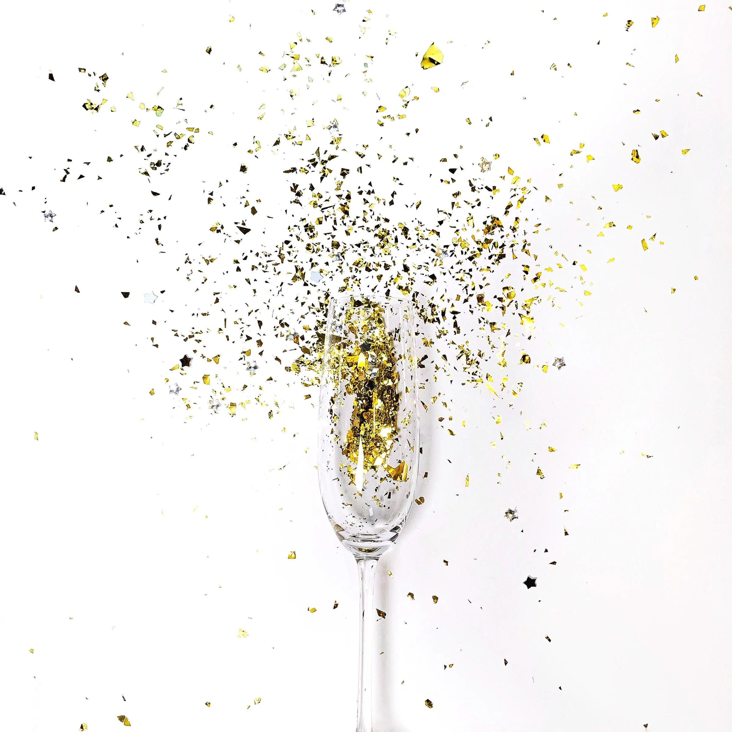 A champagne flute glass filled with champagne, surrounded by gold and black confetti, with confetti bursting out above the glass against a white background.