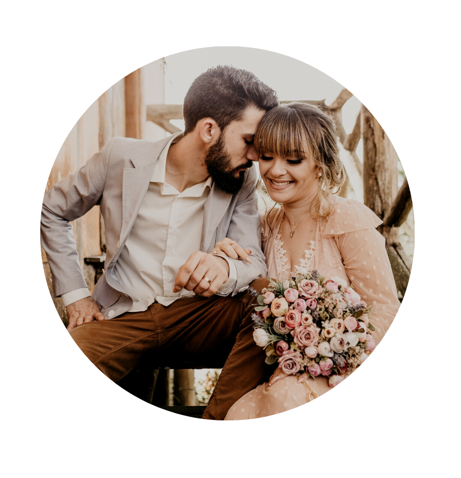 A man and woman sharing an intimate moment, with their foreheads touching. The woman is holding a large bouquet of pink and white flowers, and they are sitting on rustic wooden stairs in a cozy setting.
