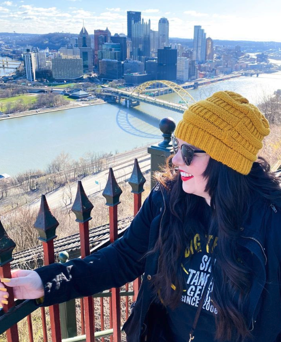 Woman in yellow knit hat and sunglasses smiling, with a city skyline and river in the background.
