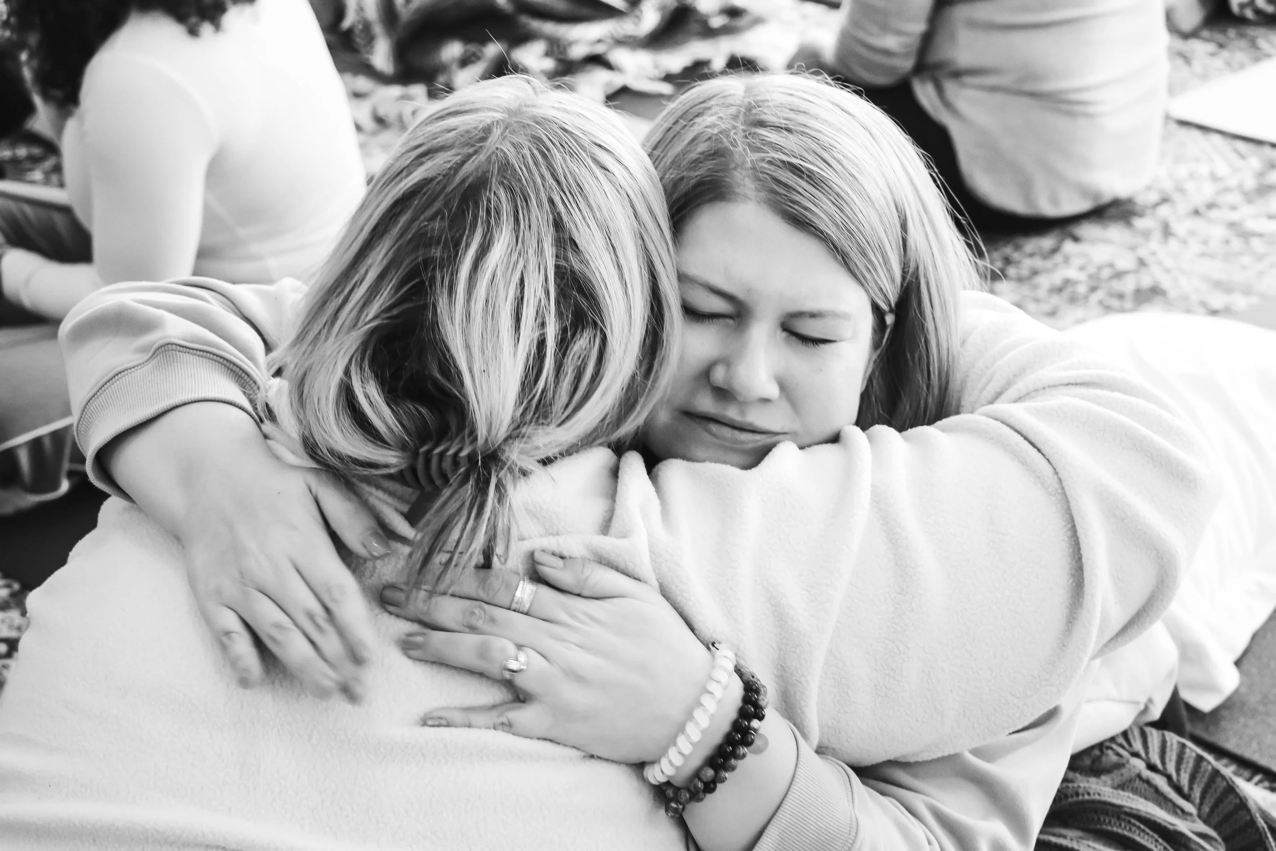 Two women hugging at a group gathering, one with eyes closed, expressing emotion in black and white.