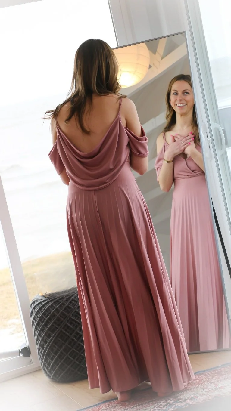 Woman in a pink dress looking at her reflection in a mirror, standing indoors.