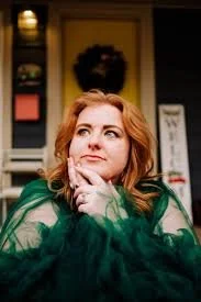 Woman with red hair in green clothing, hand on chin, sitting outdoors, thinking