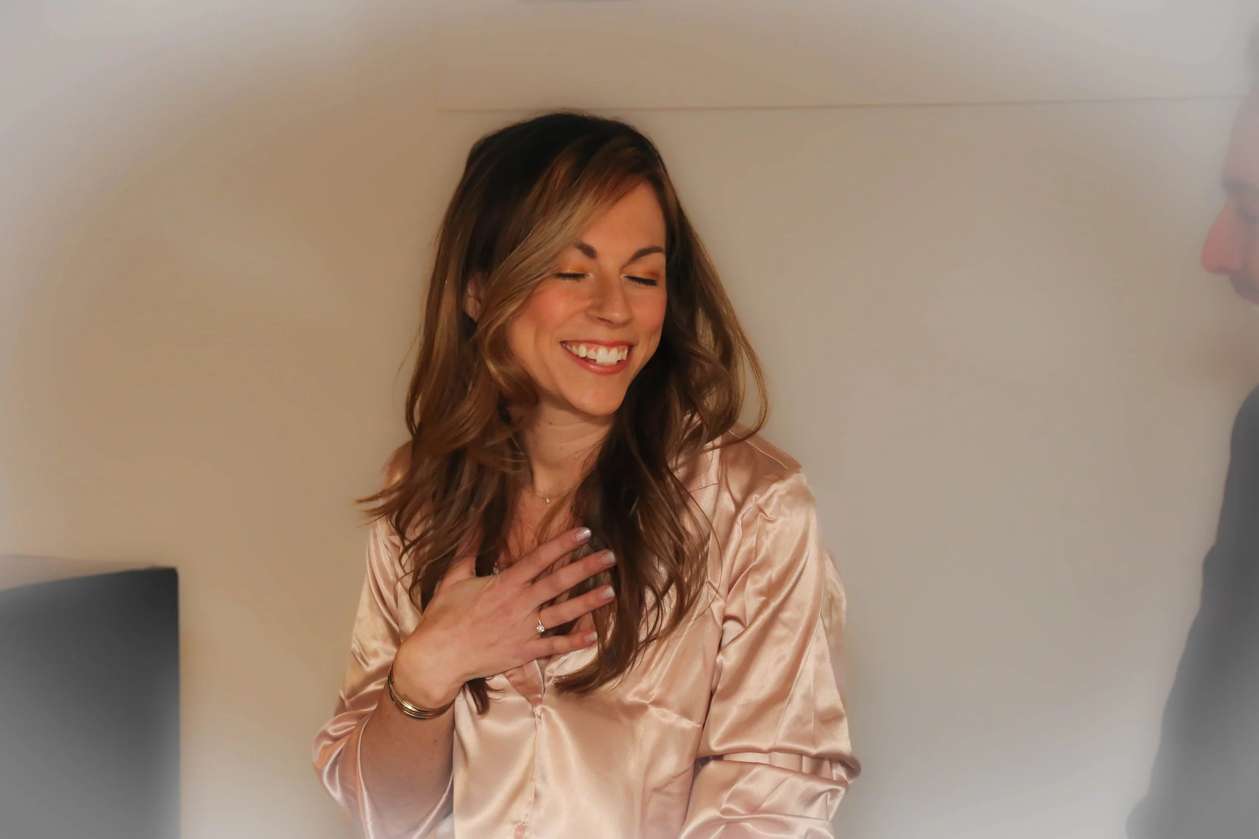 Woman smiling with eyes closed, wearing a satin shirt, hand on chest.