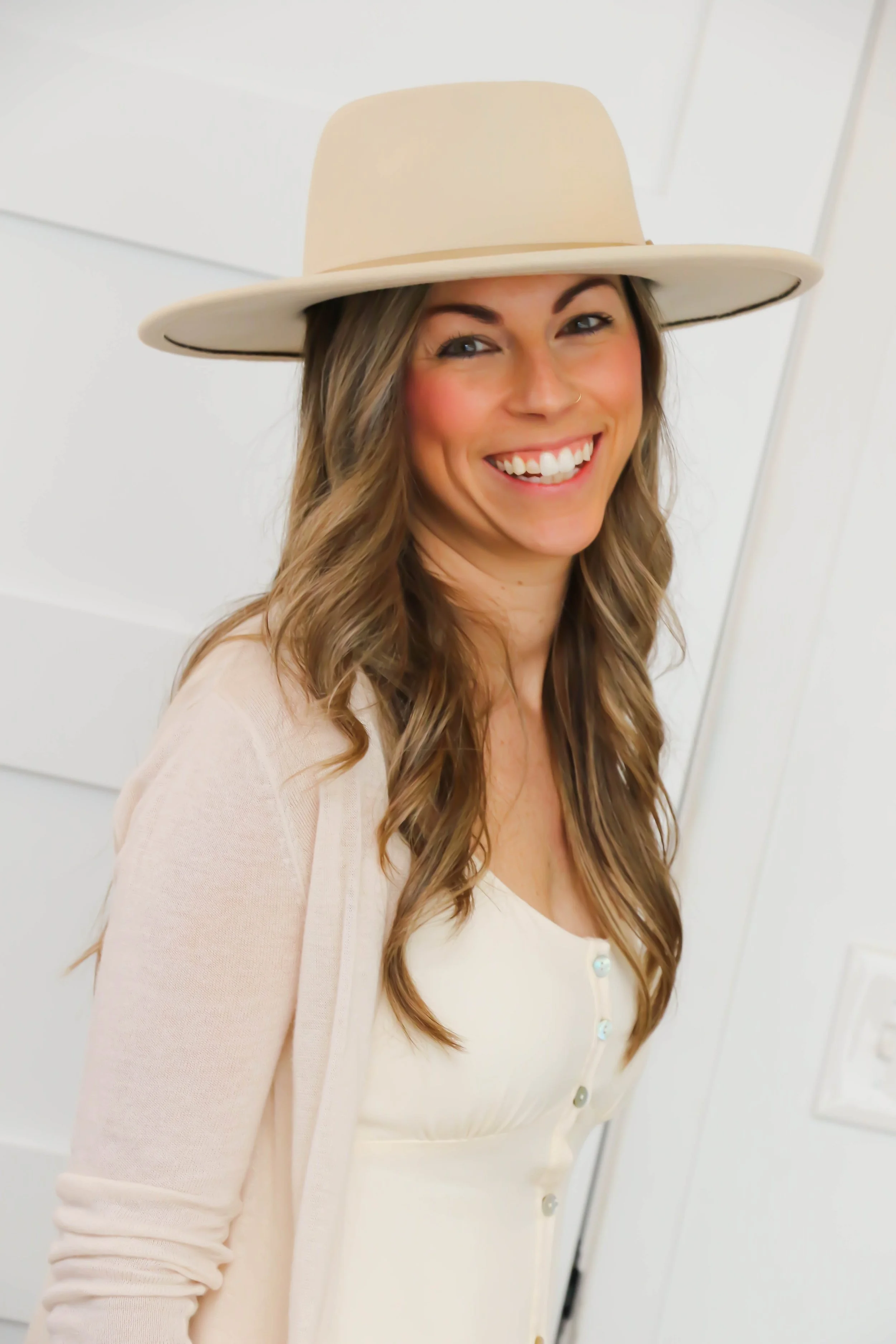 Woman smiling, wearing a beige hat and cardigan in a light-colored room.