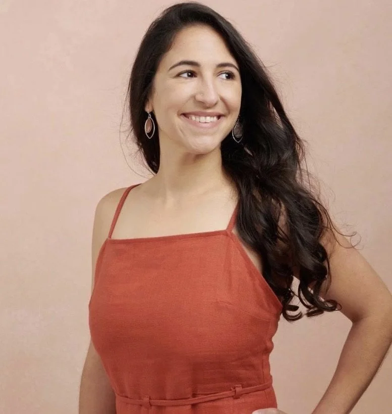 Smiling woman in an orange dress with long dark hair against a beige background.