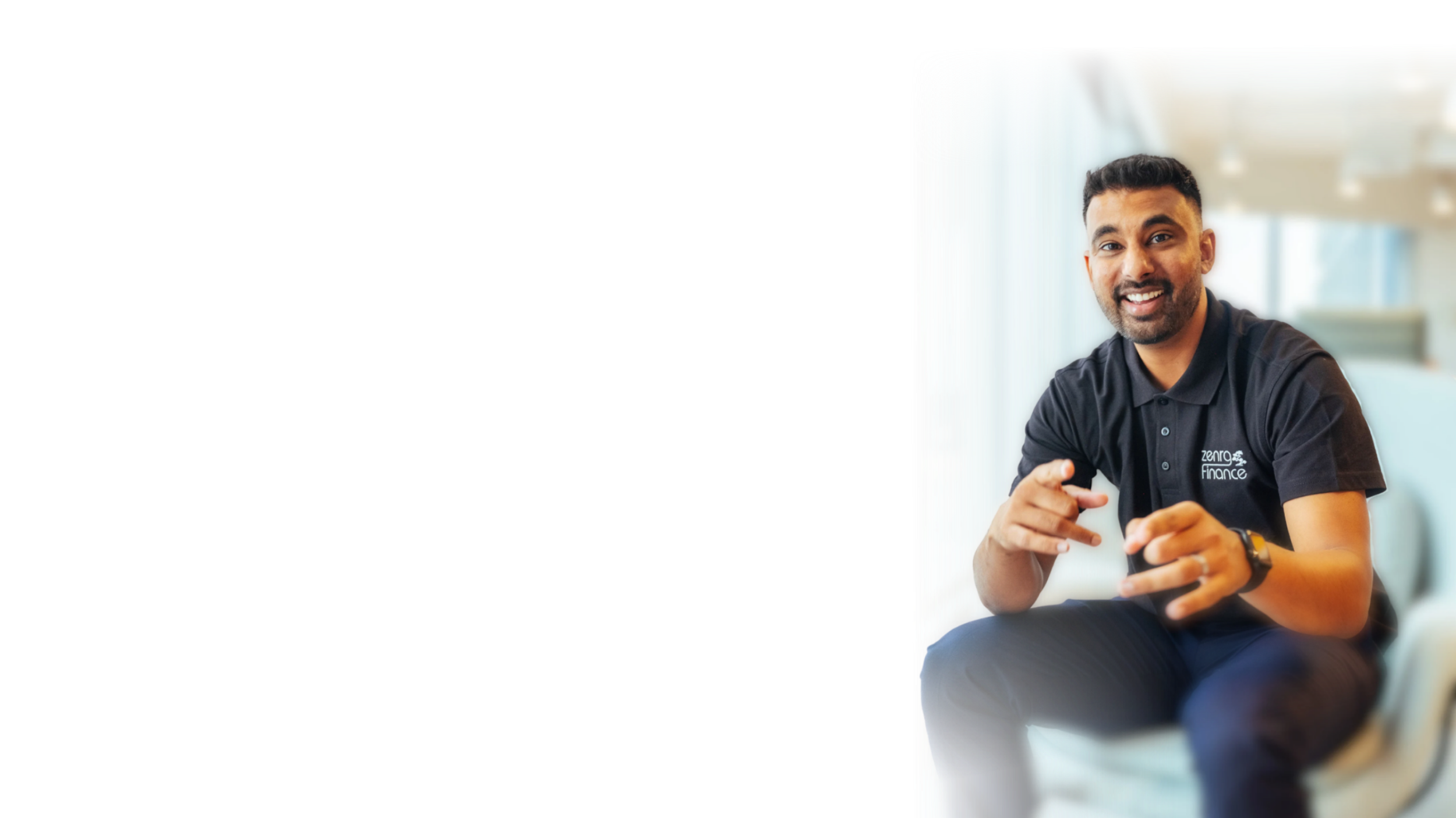 A man with short black hair, a beard, and wearing a black polo shirt with a logo that says 'Zero Finance' is smiling and pointing at the camera while sitting indoors with a blurred background of windows.