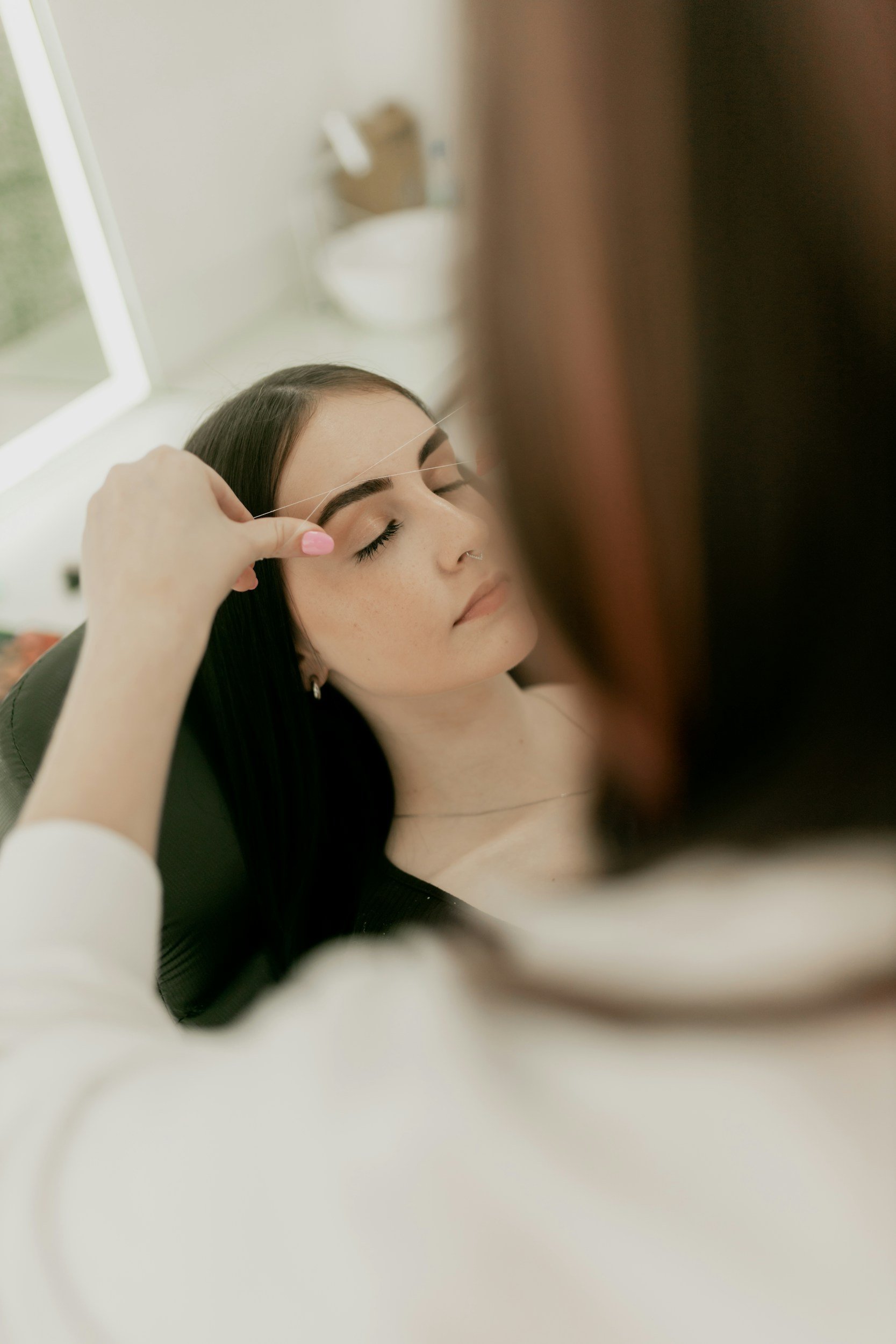 A woman with closed eyes receiving eyebrow threading service, with a person in the foreground placing a thin thread into the eyebrows.