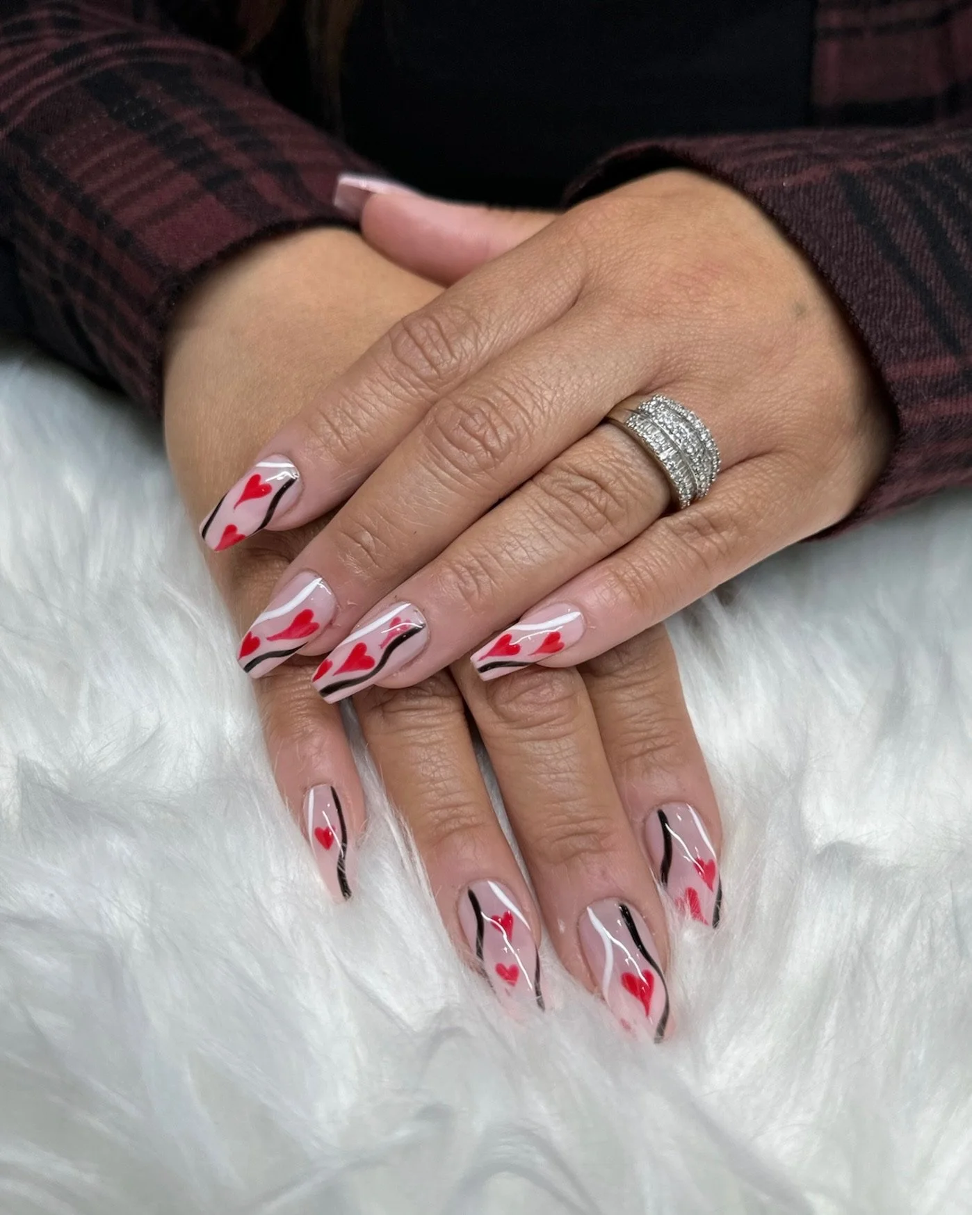 Close-up of a person with manicured nails featuring a Valentine's Day theme, including red hearts and black wavy lines on a white and natural base, with a silver ring on the ring finger, resting on a white fuzzy surface, wearing a burgundy plaid shirt.