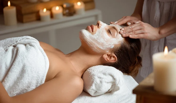 A woman receives a facial treatment with a white facial mask in a spa, candles in the background, and a relaxing atmosphere.