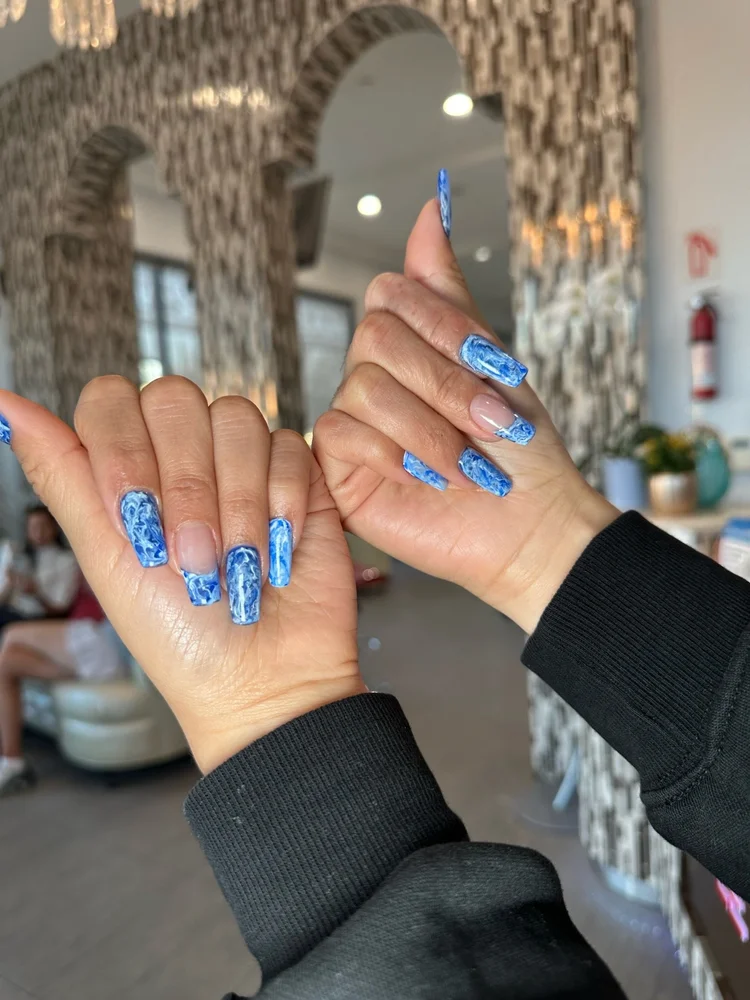 Close-up of hands with blue marble-patterned gel X nails, indoors with a blurred background of modern interior decor and a person seated in the background.