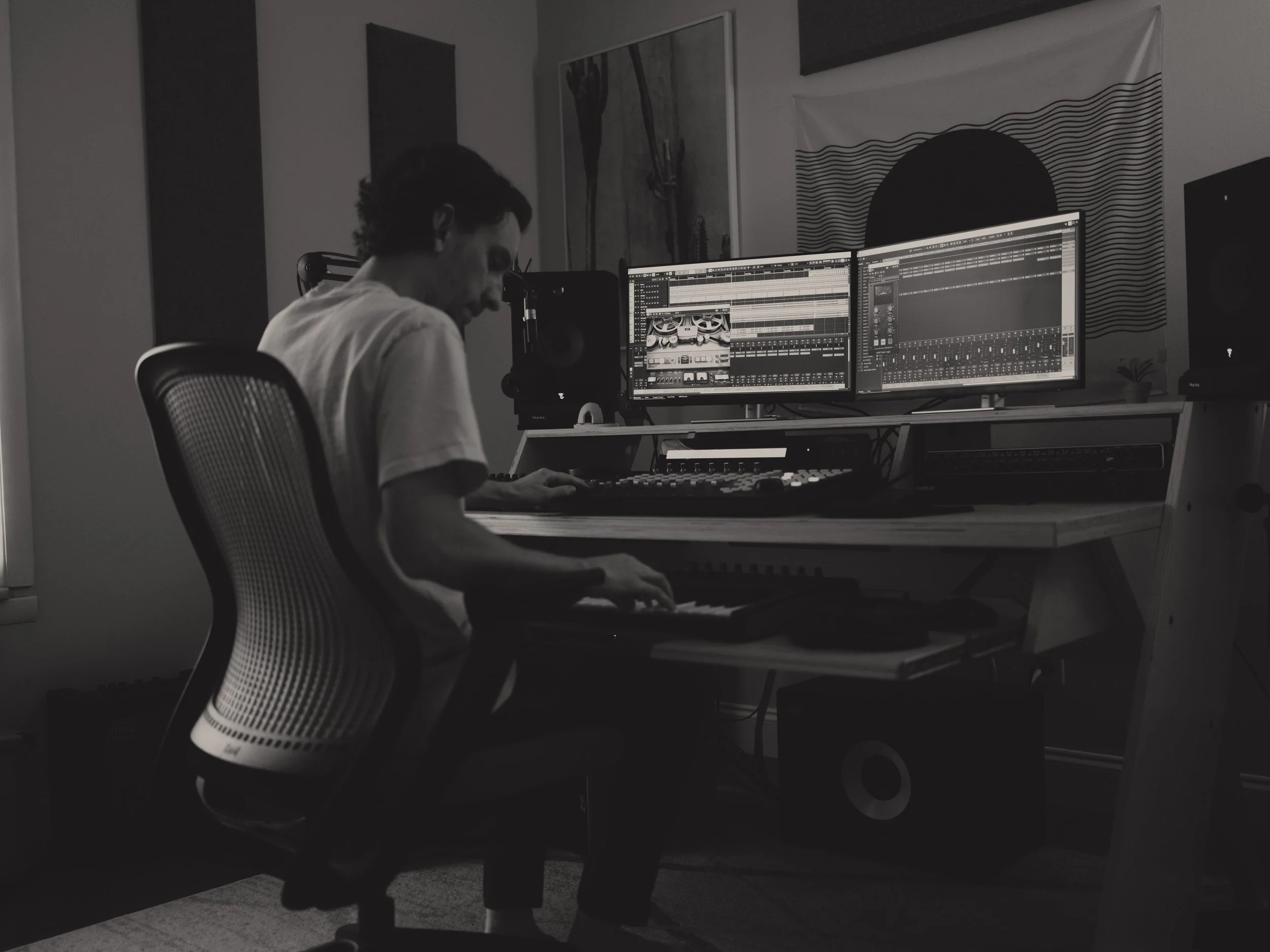 Person working at a music production computer station with dual monitors, mixing console, and studio speakers.