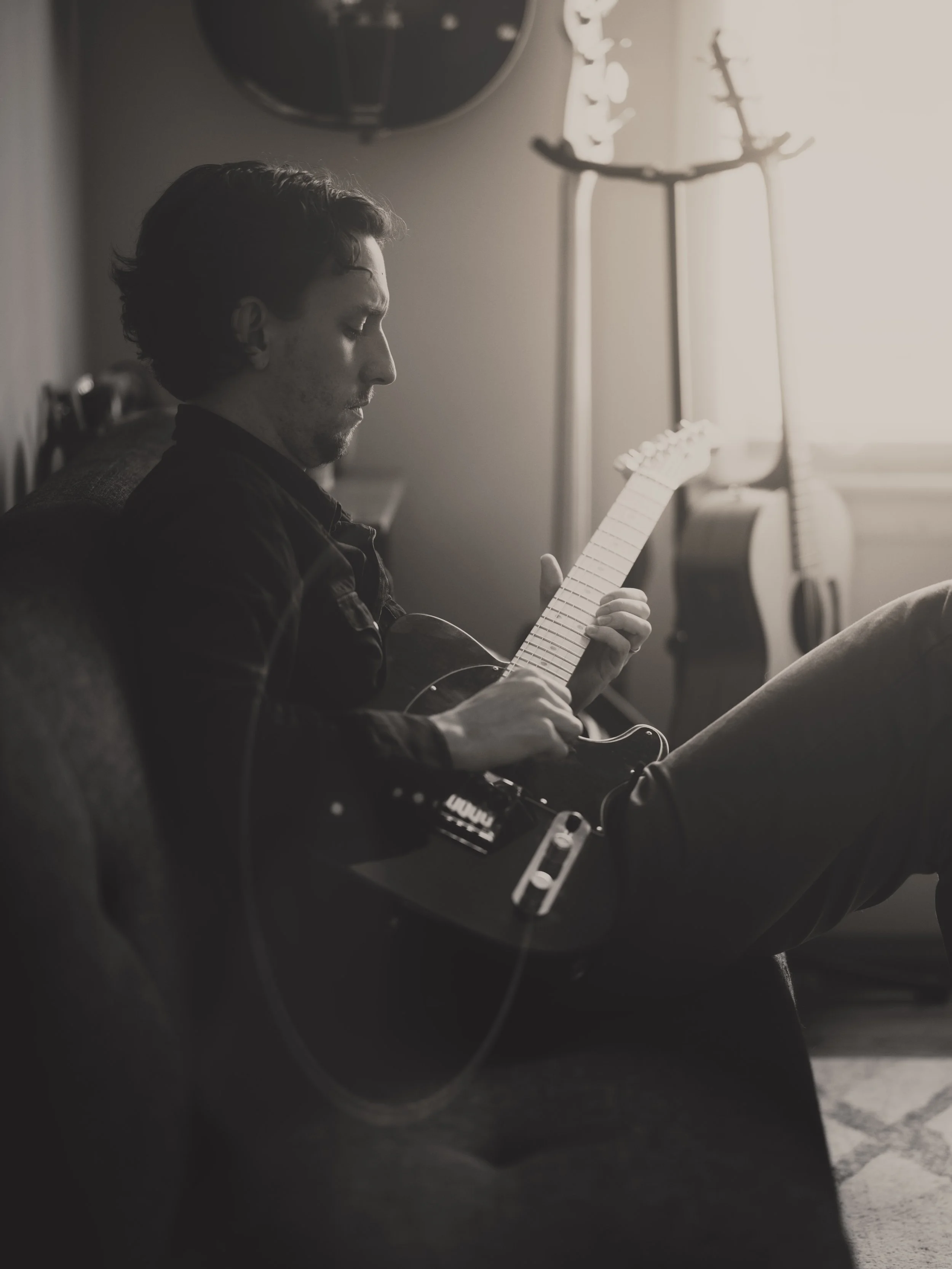 A man sitting on a couch playing an electric guitar in a sunlit room, with two acoustic guitars hanging on the wall behind him.