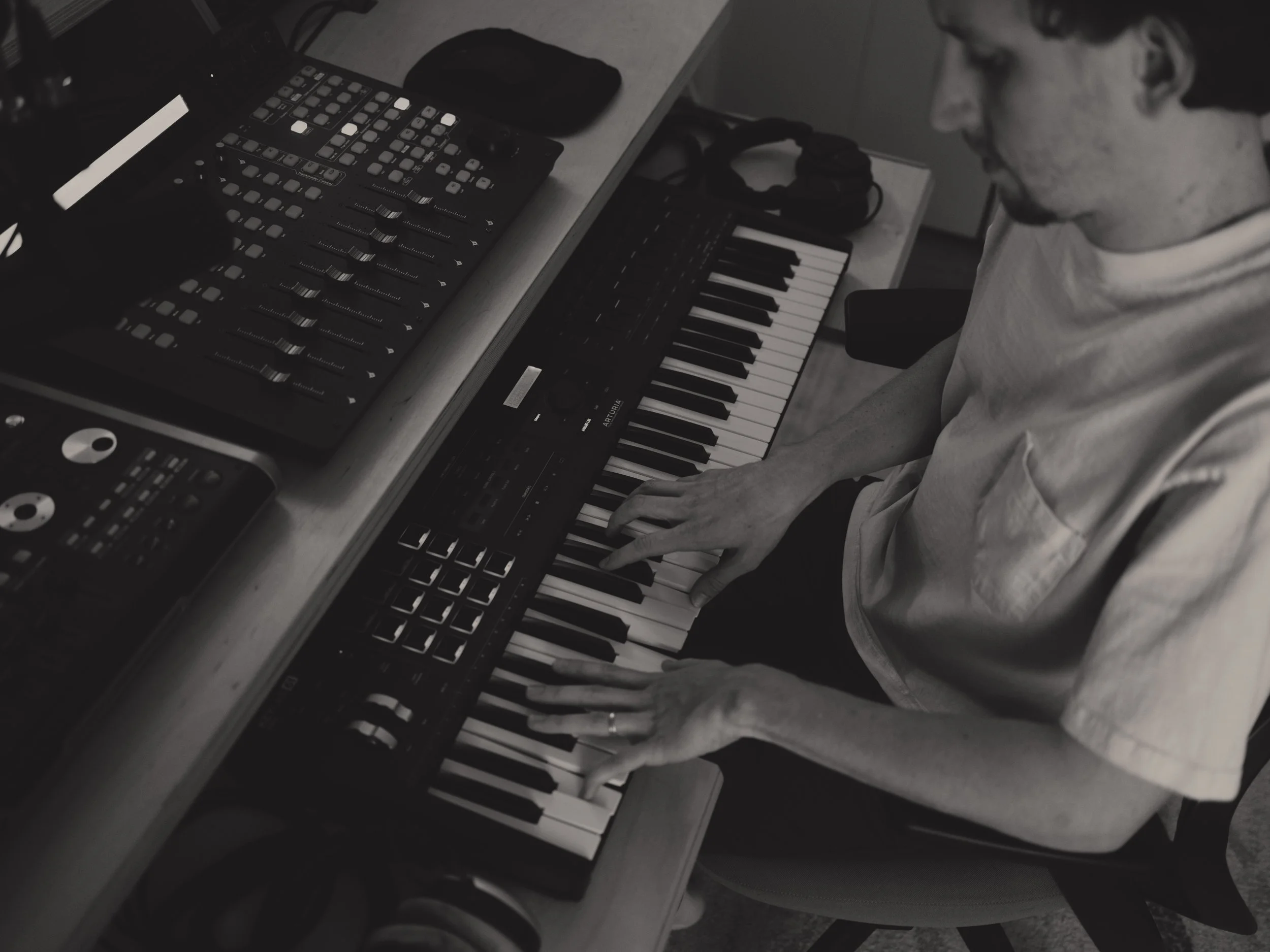 Person playing keyboard in a music studio, with electronic music equipment and synthesizers on the desk.