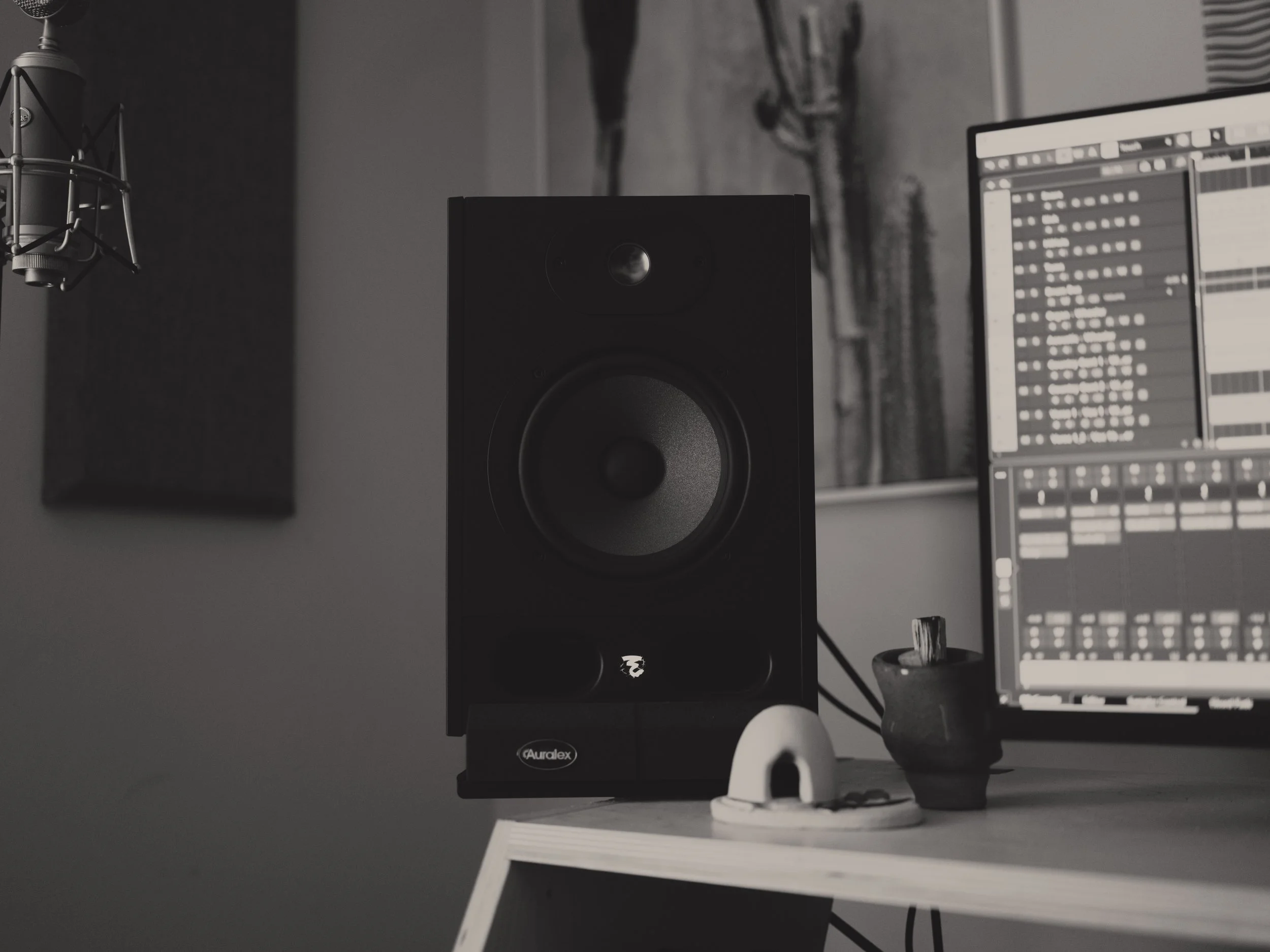 A music studio workspace with a black studio monitor speaker, a computer monitor showing digital audio workstation software, a microphone, and some small decorative items on a wooden desk in a gray-walled room.