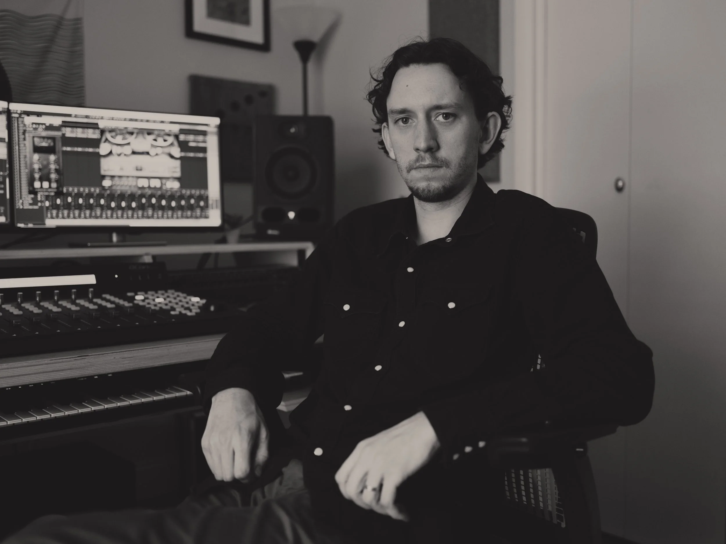 A man with curly hair and a serious expression sitting in a music studio with a computer, speaker, and keyboard in the background