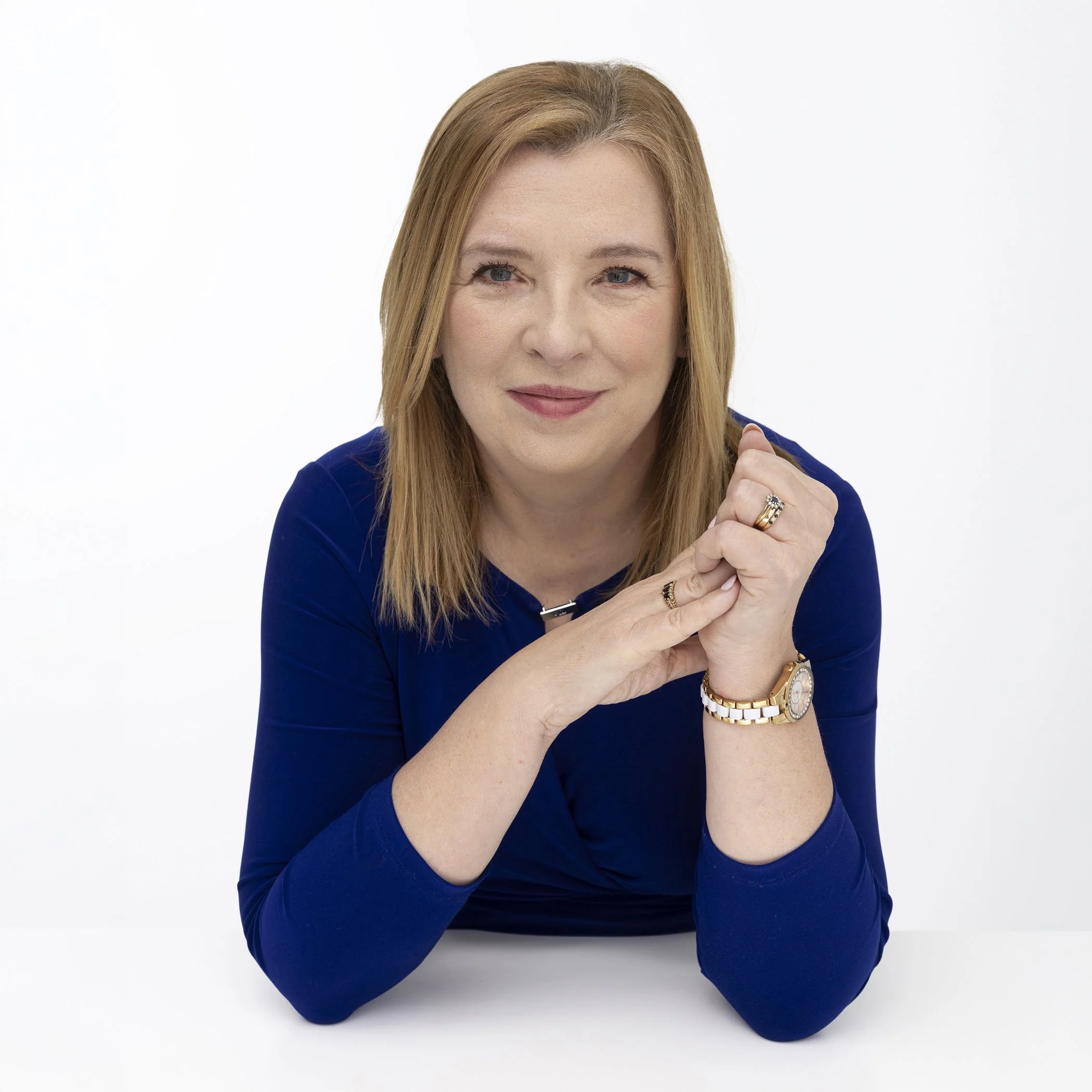 A woman with blonde hair and blue eyes smiling, wearing a blue top, resting her chin on her clasped hands, accessorized with rings and a gold watch, against a white background.