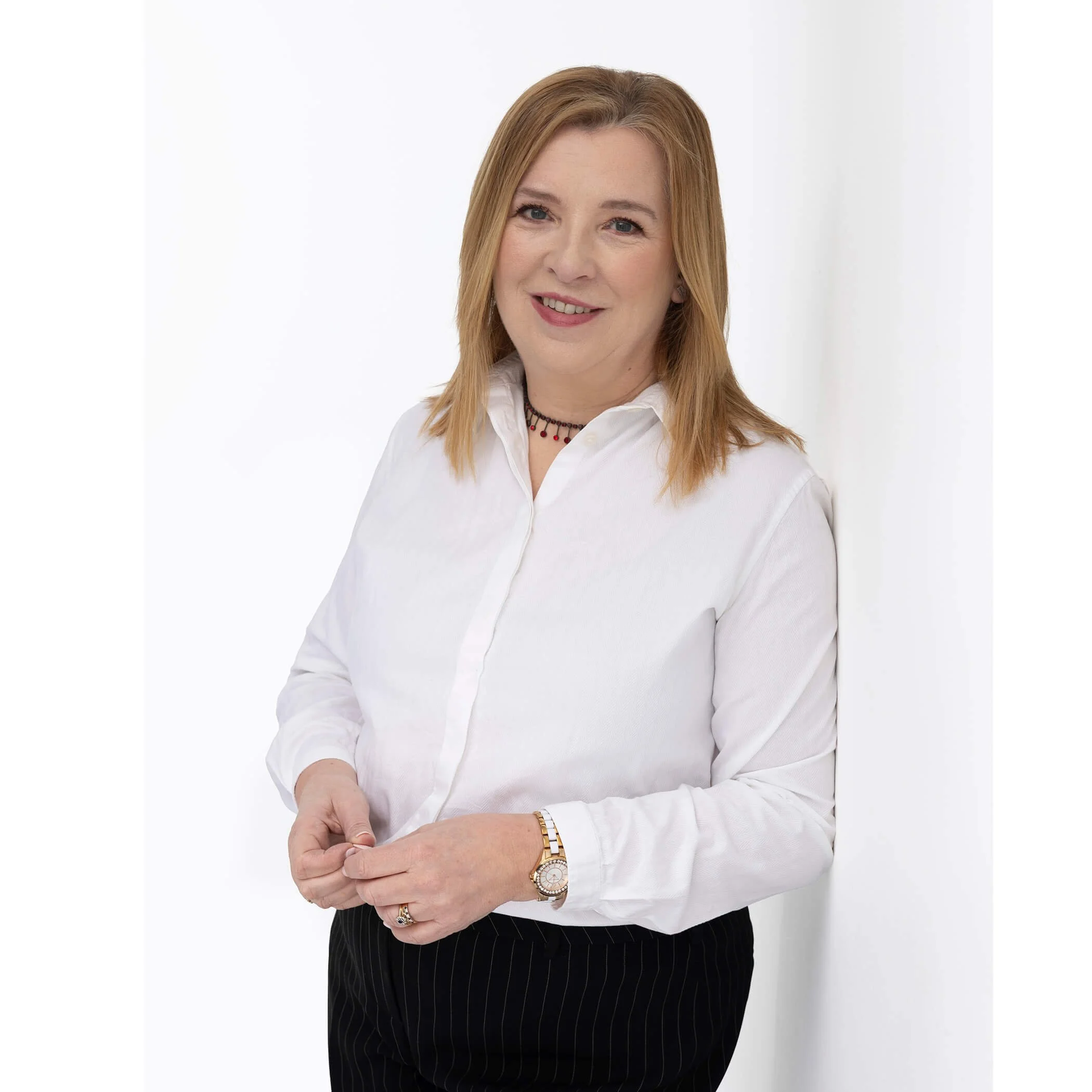 Maureen McCowen in a white shirt, standing with hands lightly clasped, smiling softly against a white background.