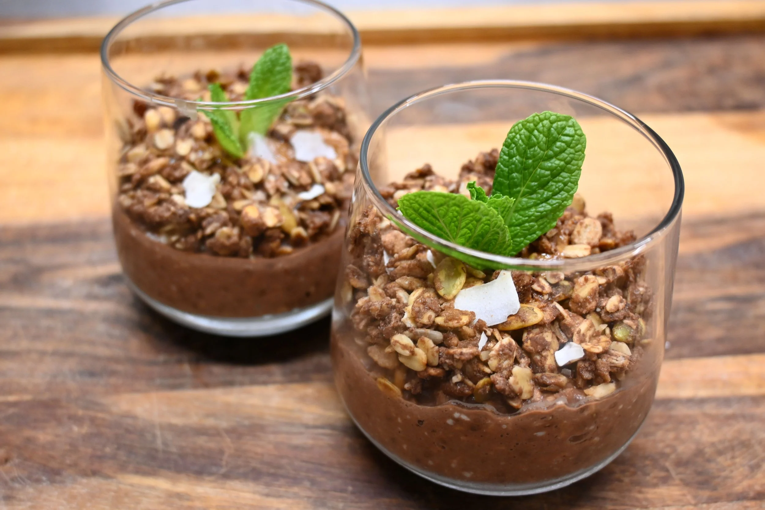 Chocolate mousse topped with granola and mint leaves in clear glass cups on a wooden surface.