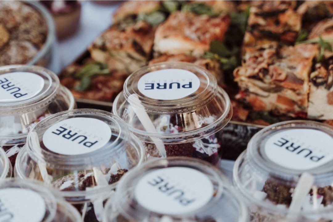 Clear plastic cups with white labels reading 'PURE' in front of a tray of sliced cold cuts or sandwiches.