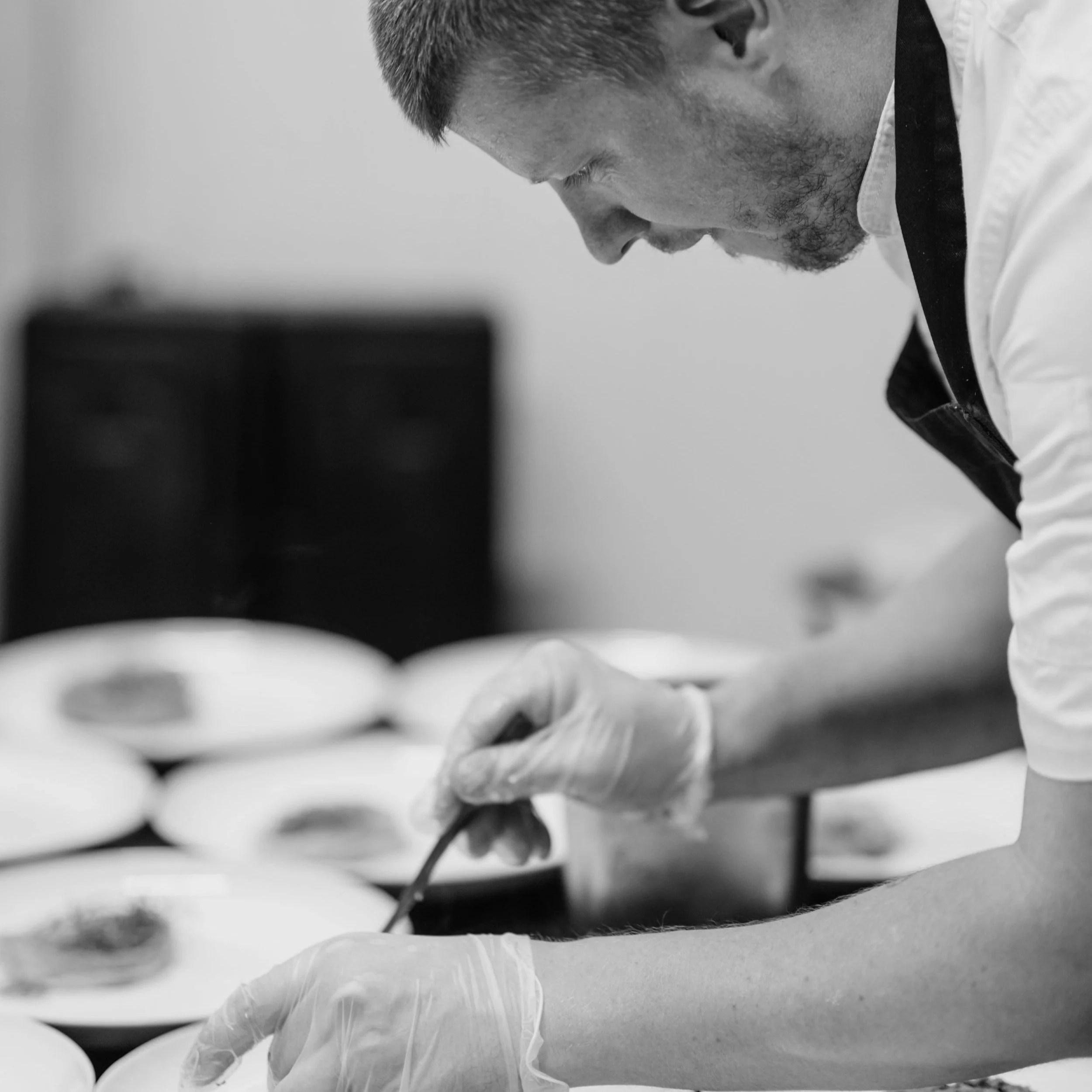 A man wearing gloves and a white shirt with an apron is preparing food with a small utensil, surrounded by plates of food.