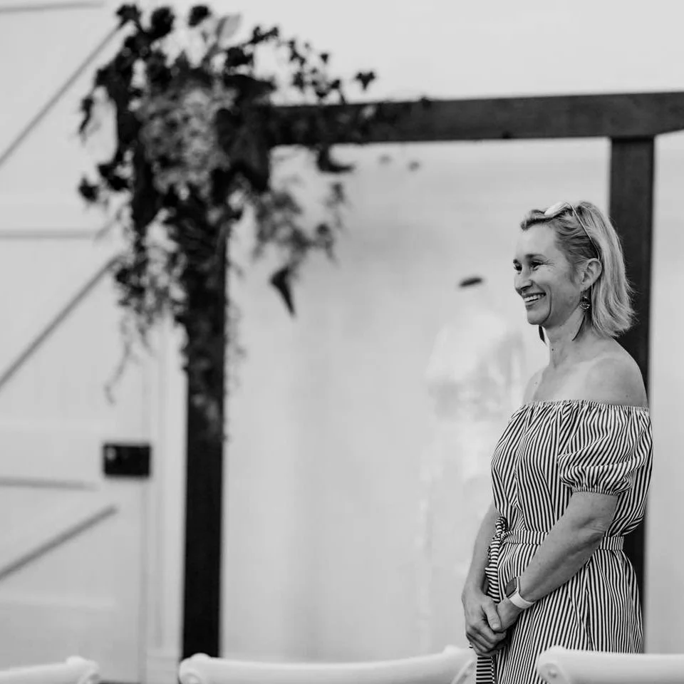 A woman in a striped off-the-shoulder dress smiling and standing near a wall at an indoor event.